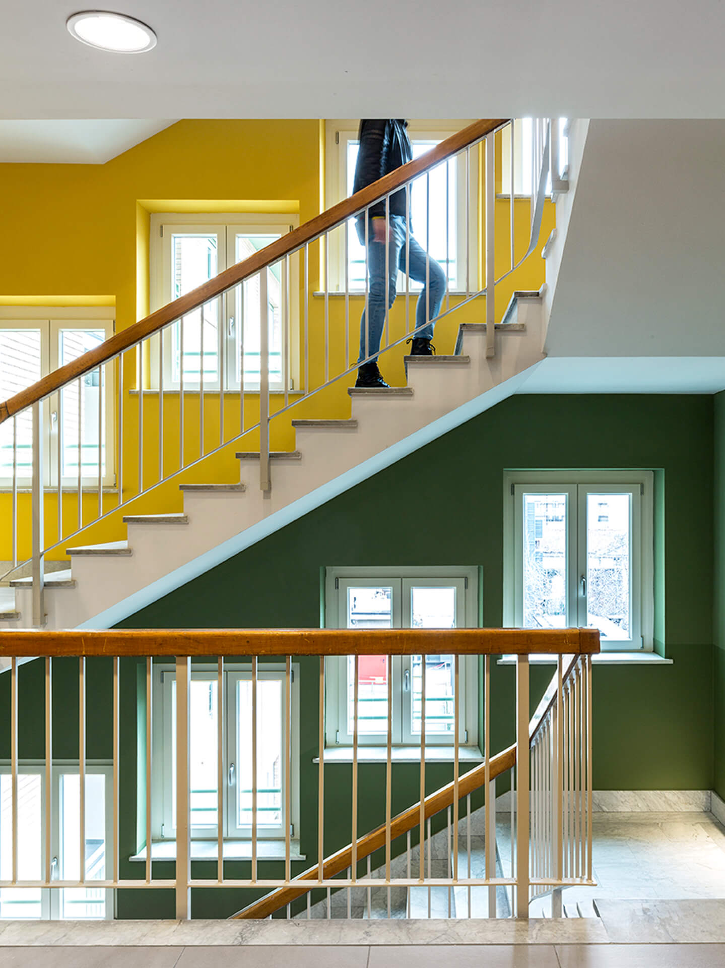 Scala interna di un edificio con pareti colorate giallo e verde, finestre bianche che illuminano lo spazio e ringhiere in metallo con corrimano in legno; una persona sta salendo i gradini tra i diversi livelli della scala.