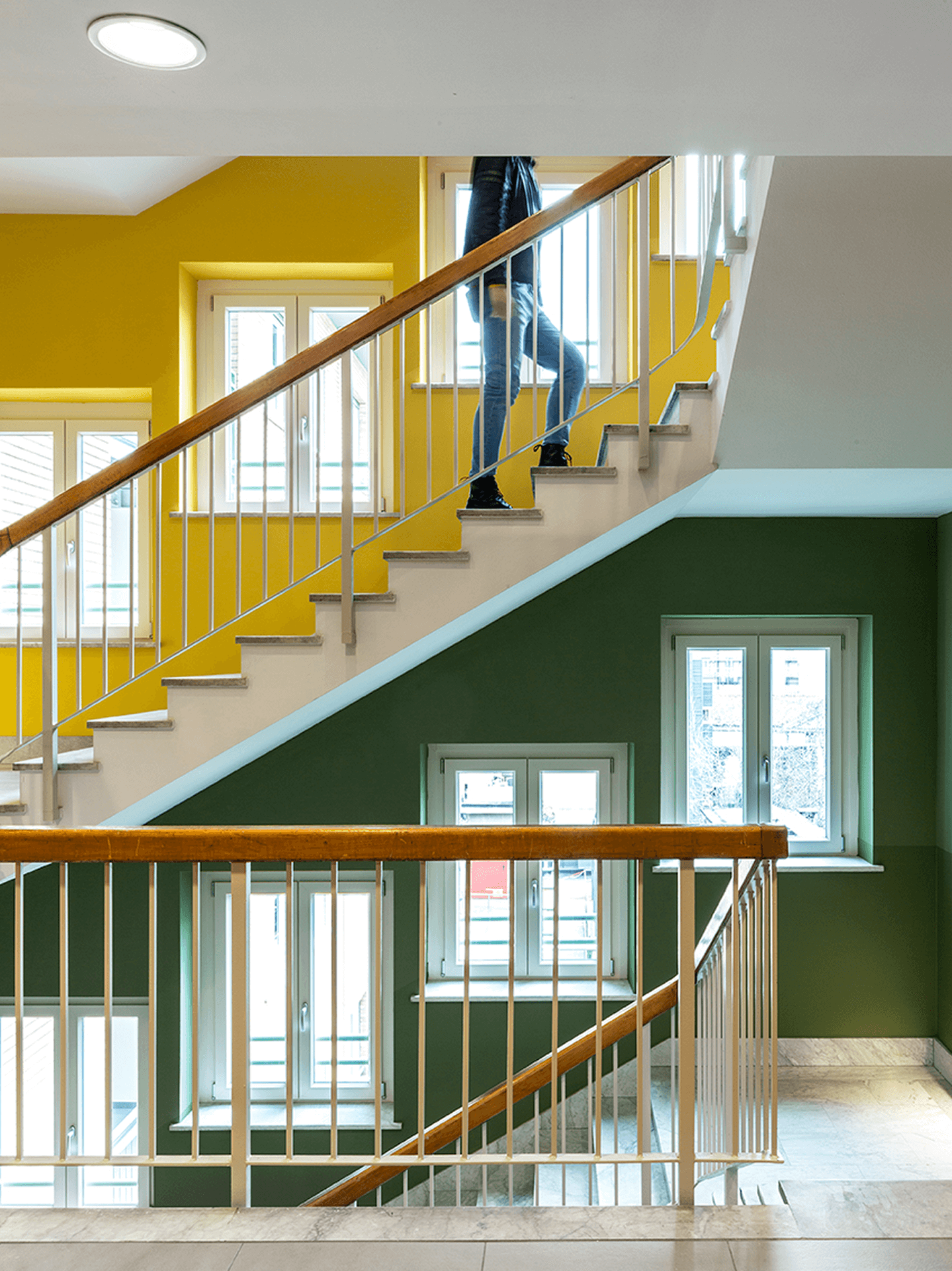 Scala interna di un edificio con pareti colorate giallo e verde, finestre bianche che illuminano lo spazio e ringhiere in metallo con corrimano in legno; una persona sta salendo i gradini tra i diversi livelli della scala.