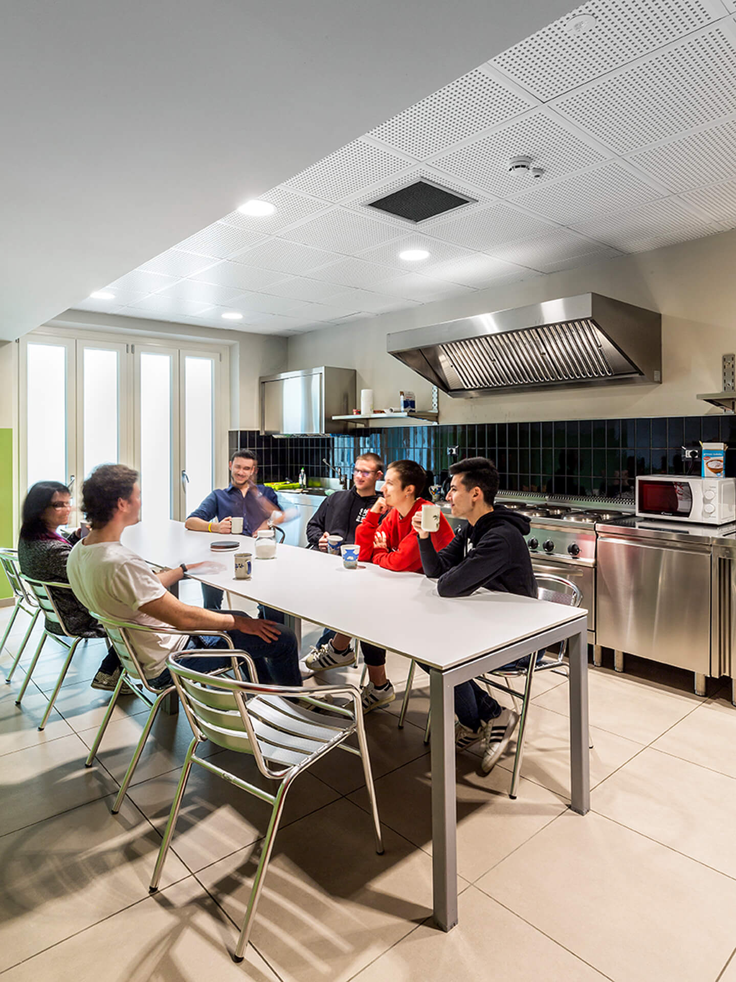 A fully equipped shared kitchen, located in a renovated space, features stainless steel furnishings, a professional hood, a hob, and dark tile cladding along the work surface. At the center of the space is a shared table with metal chairs, around which people sit drinking and conversing, demonstrating the space's convivial purpose. The space is illuminated by a modular false ceiling with recessed lighting and stoneware flooring, illustrating the design of a shared space for preparing and eating meals within a renovated building.