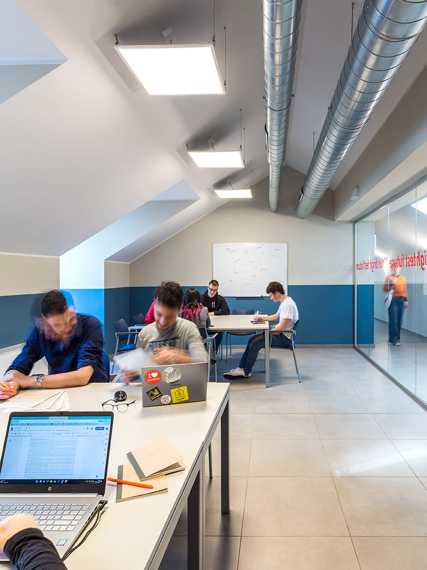 A study room created in a renovated attic, featuring a sloped roof, a partial false ceiling, and exposed utility ducts along the ridge. The space is furnished with shared tables and seating for classroom activities, used by students working with laptops and study materials. The back wall houses a whiteboard for lessons, while a glass partition separates the classroom from the corridor. The space is illuminated by suspended light panels and features a stoneware floor, demonstrating the attic's adaptation as a shared study and learning space within the renovated building.