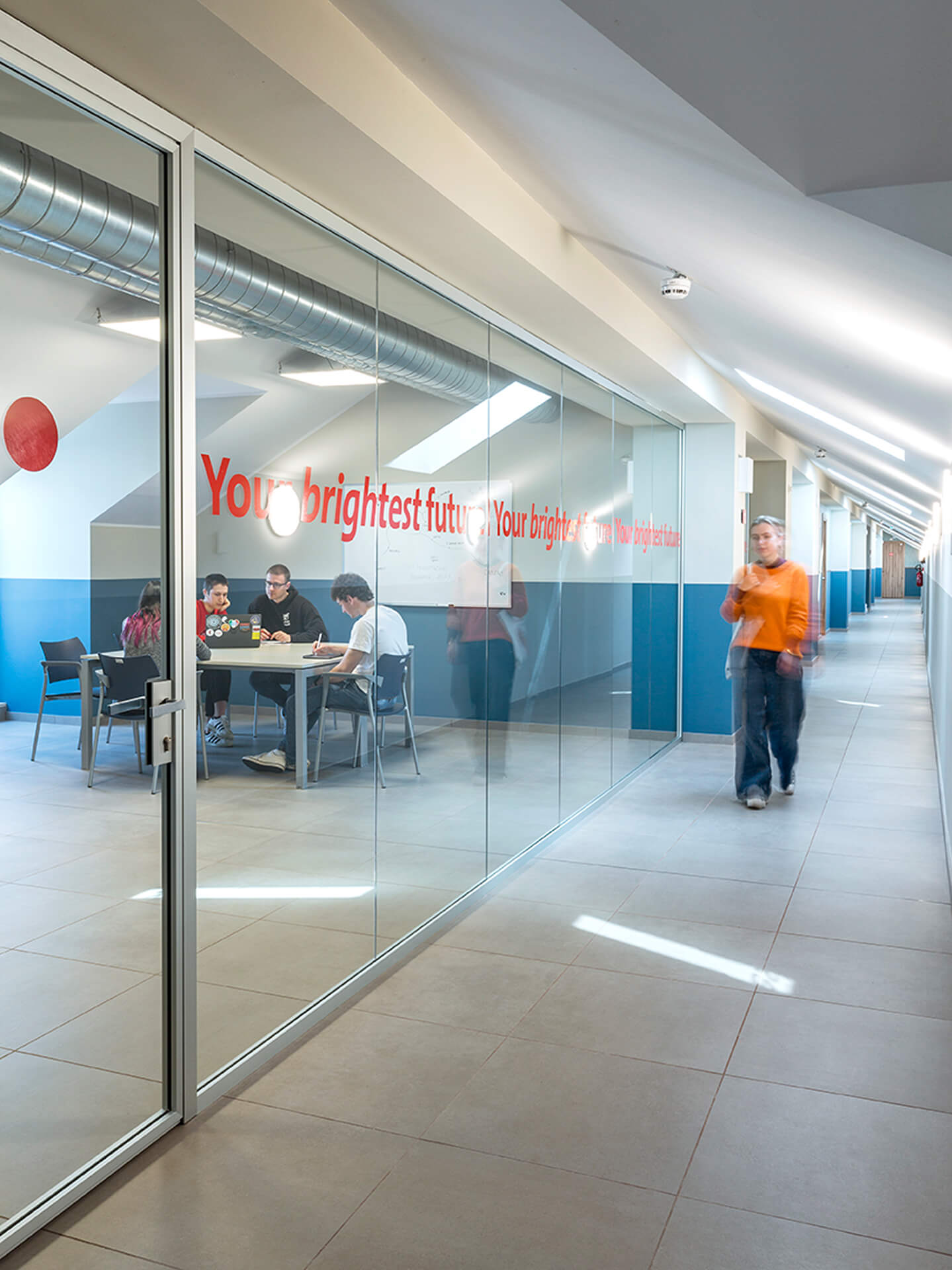 A corridor created in a renovated attic space with a sloped roof and skylights, stoneware flooring, and two-tone walls with a blue lower band. On the left side is a study room separated by a metal-framed glass wall, where several students are seated around a table working. A decorative graphic sign is applied to the glass, while a person in the corridor walks toward their room, highlighting the space's daily use. The image documents the attic's adaptation to collective and educational purposes, with contemporary finishes and transparent partitions.