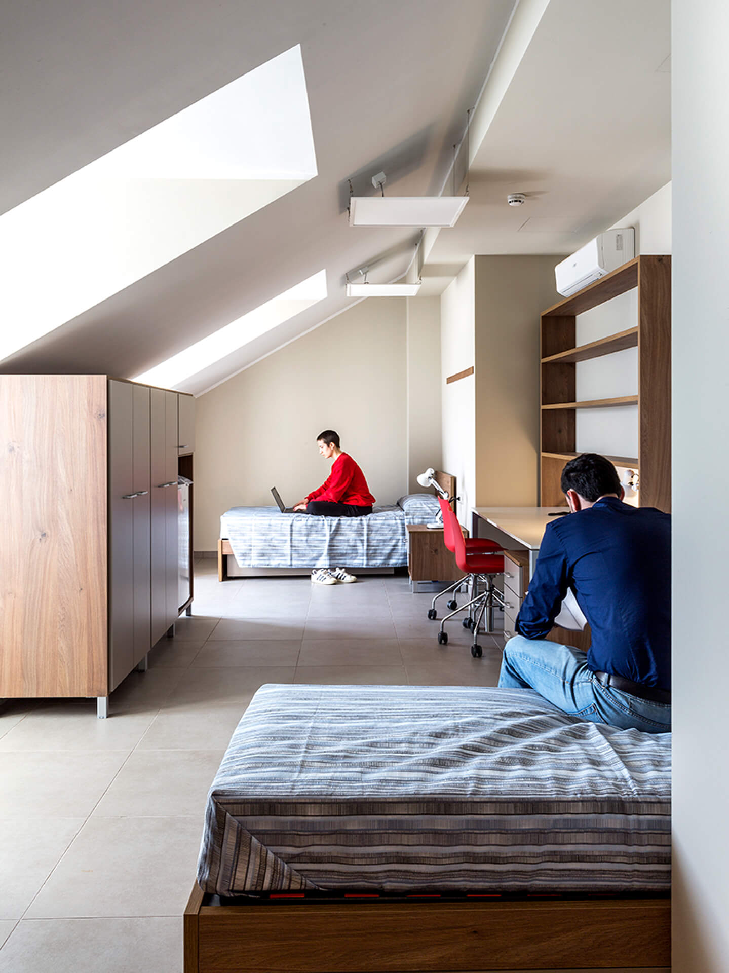 A double bedroom located in the renovated attic, featuring a sloped roof with skylights that naturally illuminate the space. The room is furnished with two single beds, wooden wardrobes, and desks with built-in seating and shelves, creating a student residential space. Two people use the room for study activities with laptops, highlighting its residential and work functions. The space features stoneware flooring, suspended lighting fixtures, and wall-mounted air conditioning, demonstrating the attic's adaptation to provide shared living space within a renovated building.