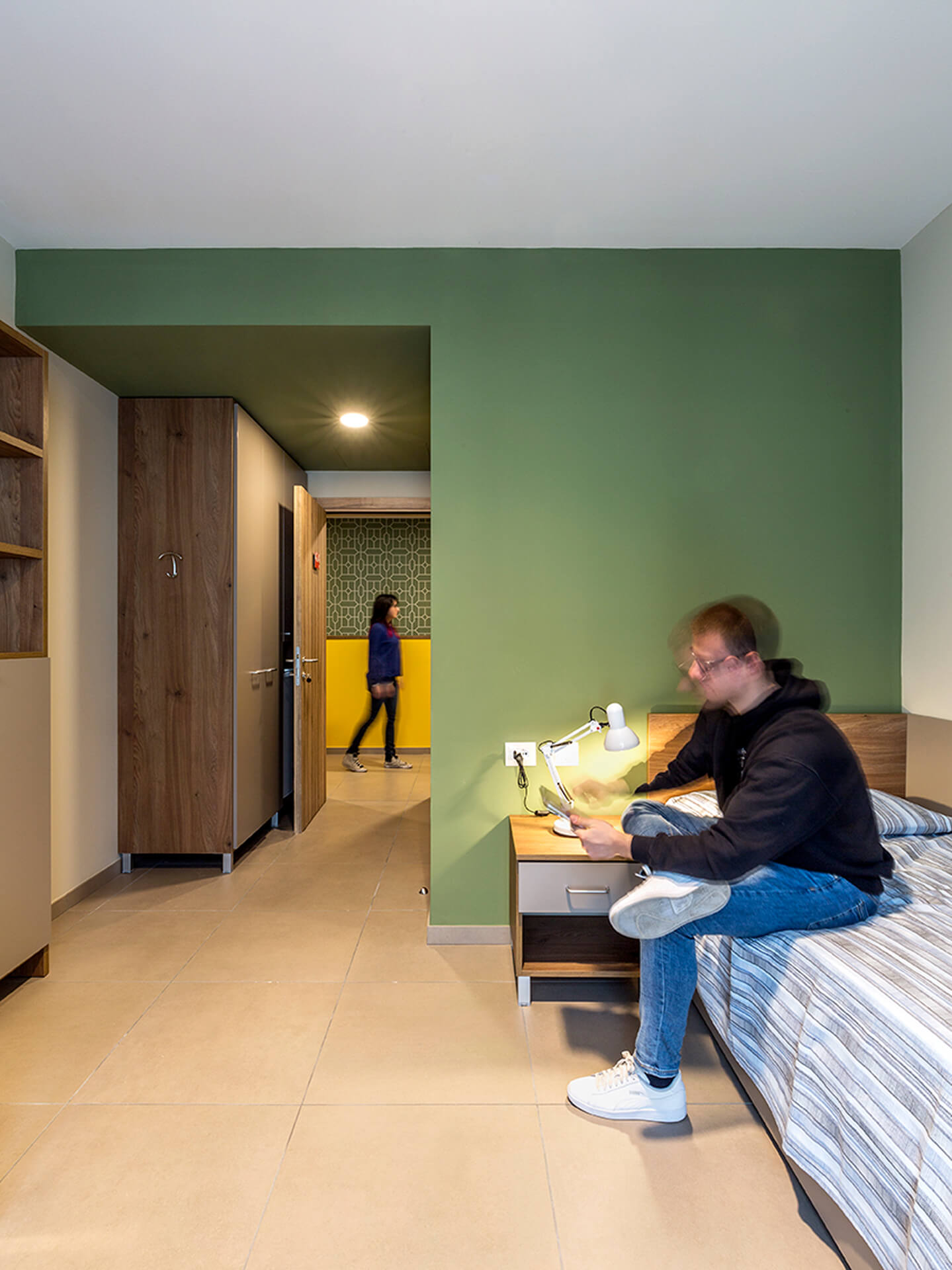 Single bedroom in a student residence in a renovated building, furnished with a bed, bedside table with lamp, wardrobe, and built-in wooden shelves. The room features stoneware flooring and a colored wall that defines the sleeping area, while in the background is the hallway leading to the shared corridor. A person is sitting on the bed using the phone, highlighting the daily use of the space, while another person walks through the corridor. The image documents the layout of essential and functional living units intended for shared student housing.