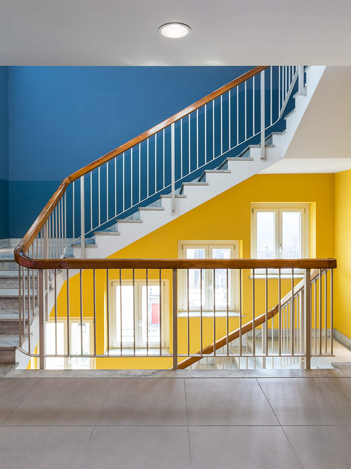 Renovated internal stairwell with overlapping ramps and painted metal parapets with wooden handrails, illuminated by windows along the perimeter wall. The surfaces are painted in different colors to identify the levels, blue at the top and yellow at the bottom, highlighting the vertical distribution of the space. The stone staircase connects the floors around a central void, while natural light from the windows enhances the renovation project with contemporary finishes within an existing building.