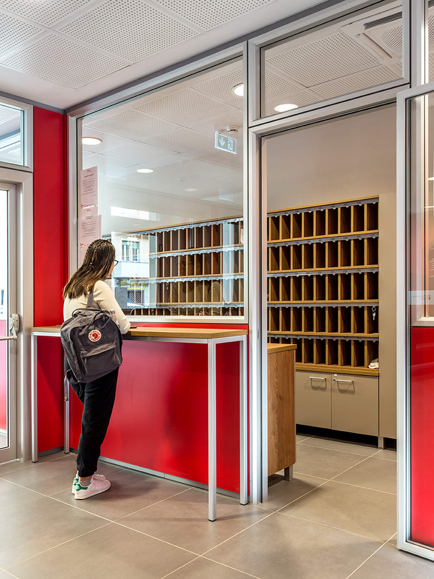 Internal reception desk with a welcome desk and glass walls made of metal profiles, featuring red paneling and contemporary furnishings. A person with a backpack leans against the desk, while in the background are shelving units for mail and key distribution. The space is illuminated by a modular false ceiling with recessed lighting and stoneware flooring, and represents a renovated entrance space with functional organization for service and reception activities.