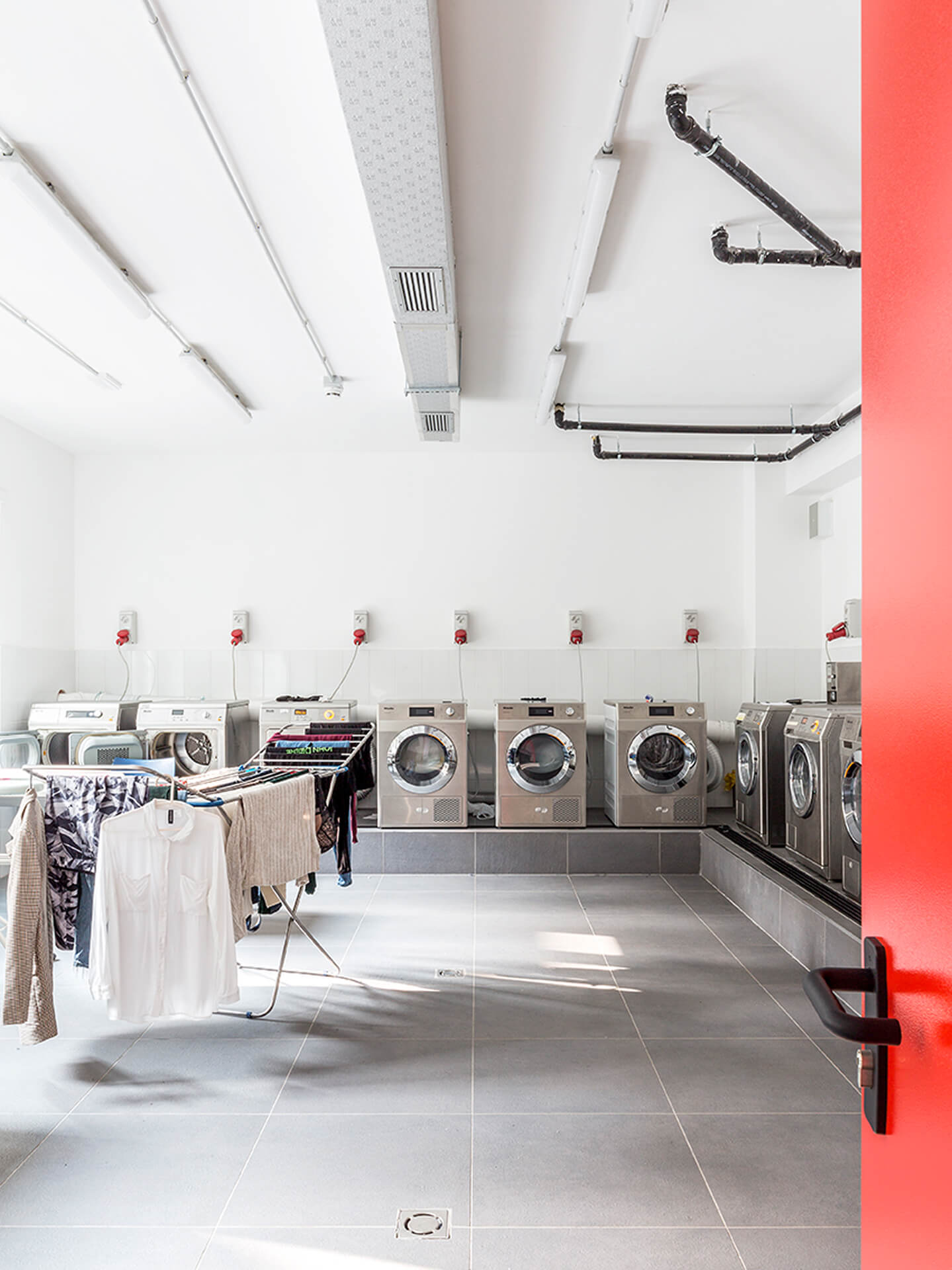 A shared laundry room in a renovated space features white plastered walls, stoneware flooring, and a ceiling with exposed wiring and linear lighting. Washing machines and dryers are lined up on pedestals along the wall, with dedicated electrical outlets and drains. Drying racks with clothes hanging out to dry are located in the center of the space, illustrating the room's intended use. The image illustrates the creation of a shared utility space within an existing building, featuring functional organization and contemporary finishes.