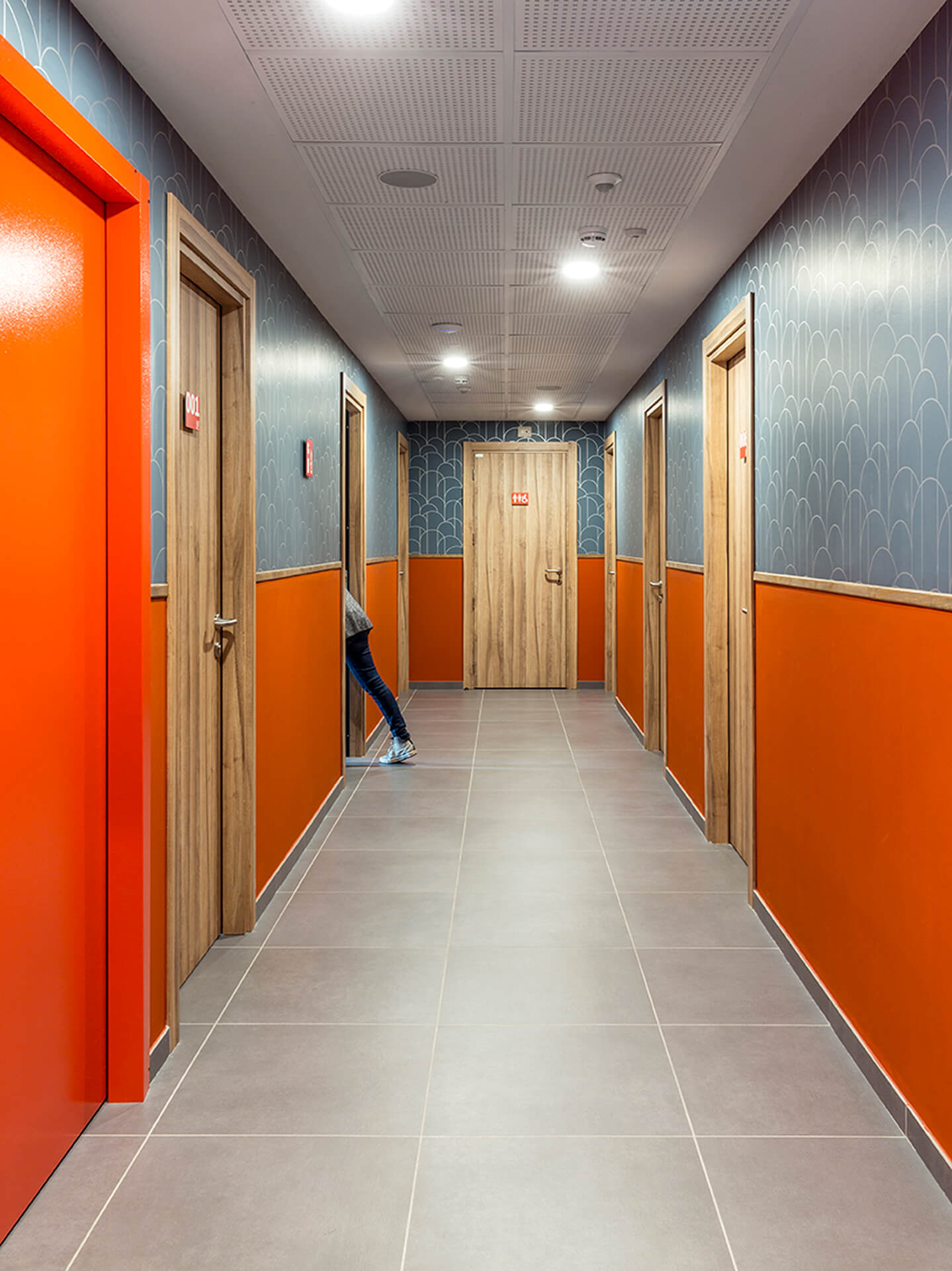 Renovated internal corridor with stoneware flooring and a modular suspended ceiling with recessed lighting, featuring two-tone orange and blue walls with decorative cladding on the upper section. Along the corridor, numbered wooden doors identify the rooms, while a person enters one of the rooms, illustrating the use of the space. The image documents the renovation of a residential or hospitality floor with contemporary finishes, distinctive colors, and functional organization of the room accesses.