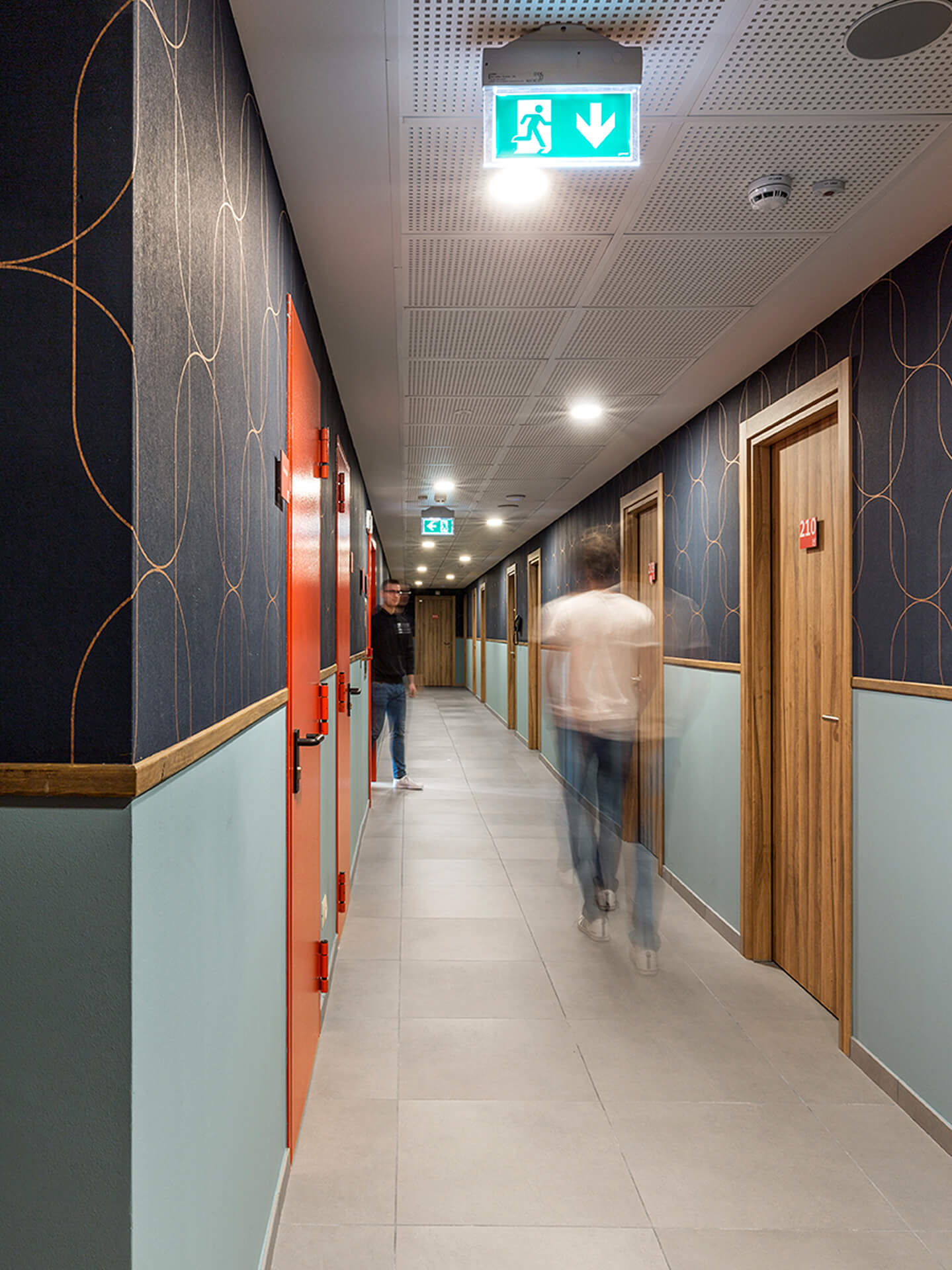 Renovated internal corridor with stoneware flooring, a modular suspended ceiling with recessed lighting, and ceiling-mounted emergency signage. The walls are finished with colored skirting and dark decorative paneling with a geometric pattern, while the corridor features numbered wooden doors identifying the rooms. Two people in motion cross the space, highlighting its daily use. The image documents the redevelopment of a residential or hospitality floor with contemporary finishes, distinctive colors, and functional organization of the internal passageways.