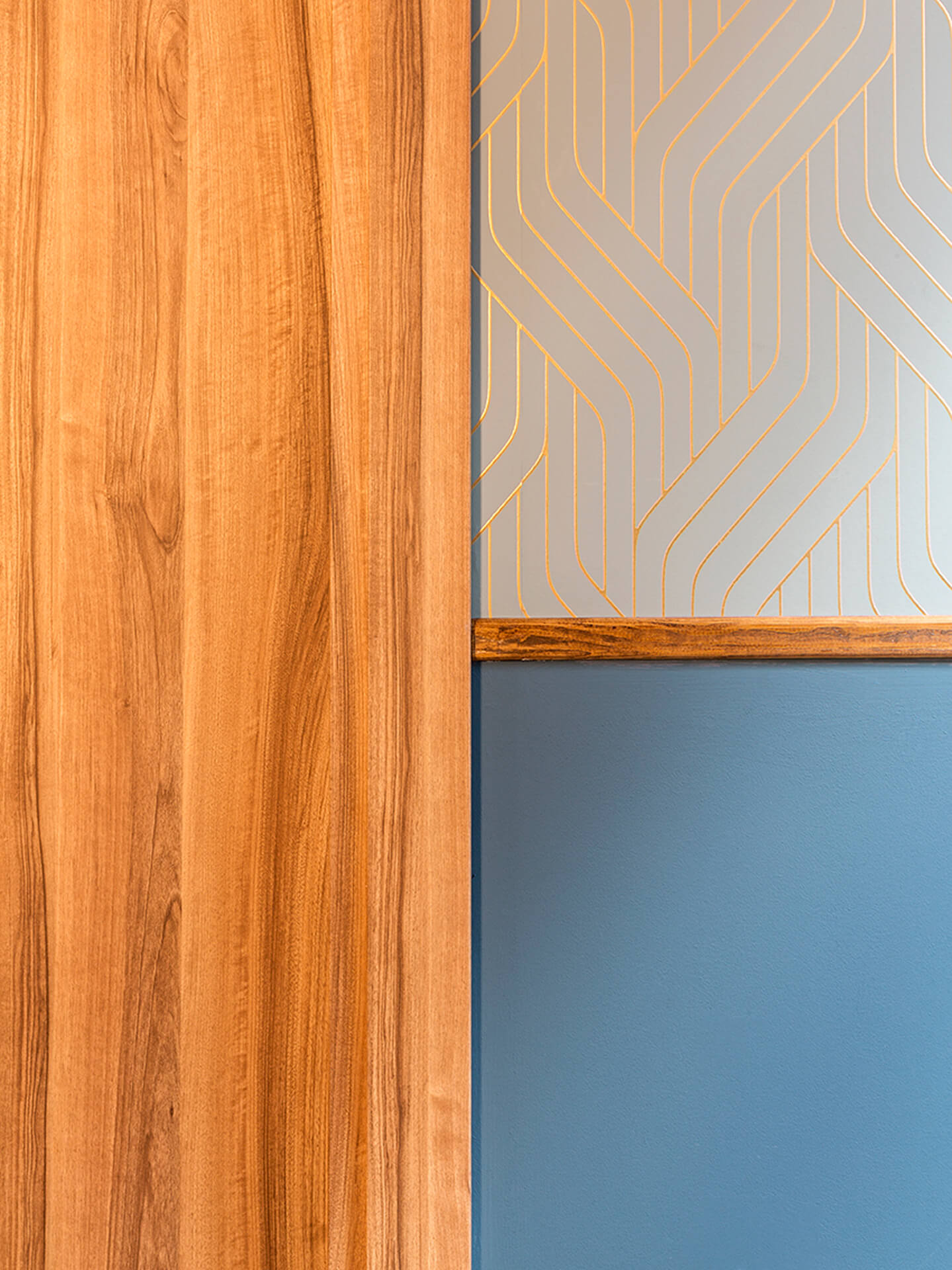 Detail of interior finishes after renovation, featuring a natural-effect wooden door and a two-tone wall composed of a lower strip painted blue and an upper strip covered with decorative geometric wallpaper. A wooden profile separates the two materials, highlighting the attention to detail and the coordinated use of colors and textures in the building's common areas. The image illustrates the choice of contemporary materials incorporated into an interior renovation project.