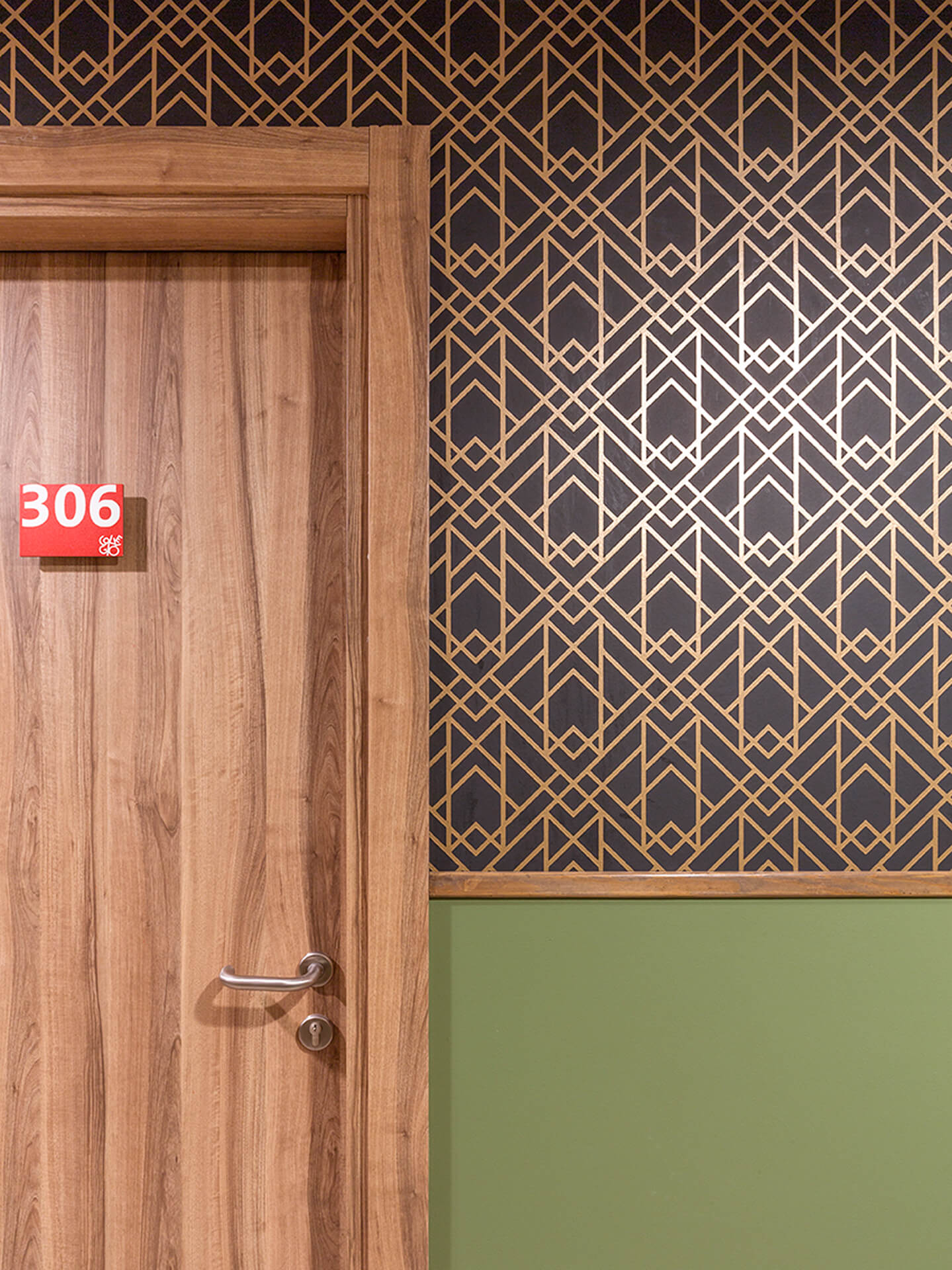 Detail of a numbered interior door with a wood-effect finish set into a decorated wall, composed of a lower strip painted green and an upper strip covered in gold geometric wallpaper on a dark background. A wooden profile separates the two materials, highlighting the meticulous attention to detail in the common areas. The room number "306" on the door identifies the layout of the apartments within a building renovated with contemporary materials and a coordinated color palette.