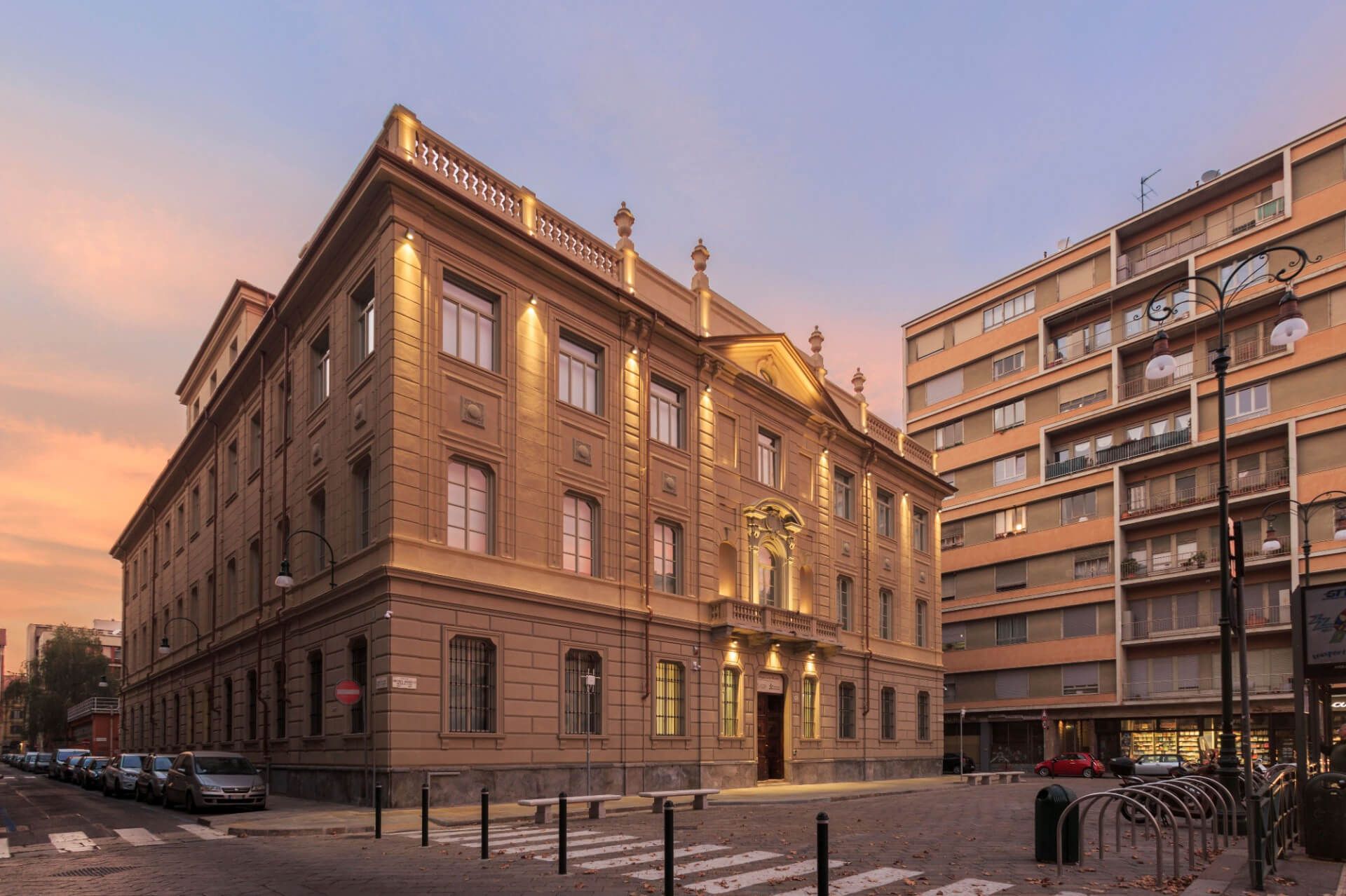 A corner building in neoclassical style, this multi-level building features a rigorously symmetrical composition, with a rusticated base and upper elevations articulated by pilasters, string courses, and molded frames around the openings. The main façade features a central axis emphasized by a monumental portal with sculptural decorations, a balcony above, and a triangular pediment, flanked by regular vertical windows. The crowning feature is a continuous balustrade interrupted by ornamental elements and pinnacles. The integrated architectural lighting, with warm, grazing light, highlights the vertical rhythm of the pilasters and the depth of the decorative details, enhancing the texture of the façade at dusk. The building is set within a diverse urban context, adjacent to modern residential buildings, with street space organized by sidewalks, pedestrian crossings, street furniture, and parking lots. The sunset sky, with its orange-pink hues, helps define the relationship between architectural volumes and light, accentuating the sculptural perception of the whole.