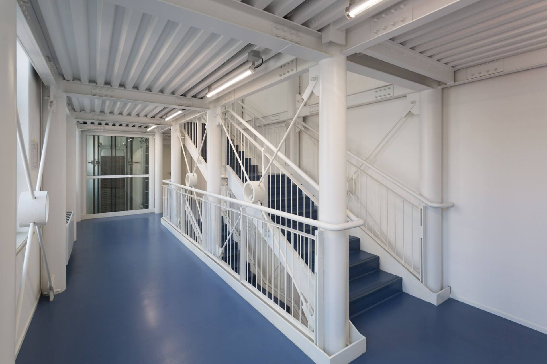 Contemporary interior space with vertical distribution and an exposed metal structure, featuring white-painted steel beams and pillars and a technical suspended ceiling with ribbed elements. The staircase, divided into multiple flights, features a metal structure with vertical bar parapets and an integrated handrail, while the steps are clad in a dark material that contrasts with the dominant white. Il pianerottolo è definito da una pavimentazione continua in resina blu, che enfatizza il carattere funzionale e industriale dell’ambiente. Elementi strutturali diagonali e nodi di connessione evidenziati contribuiscono alla lettura tecnica dell’insieme, mentre l’illuminazione lineare a soffitto garantisce una distribuzione uniforme della luce. Sullo sfondo, una chiusura vetrata delimita ulteriori spazi interni, rafforzando la trasparenza e la permeabilità visiva del progetto. L’immagine evidenzia un linguaggio architettonico industriale e contemporaneo, con particolare attenzione alla chiarezza strutturale e alla coerenza materica tra elementi portanti, finiture e sistemi impiantistici.