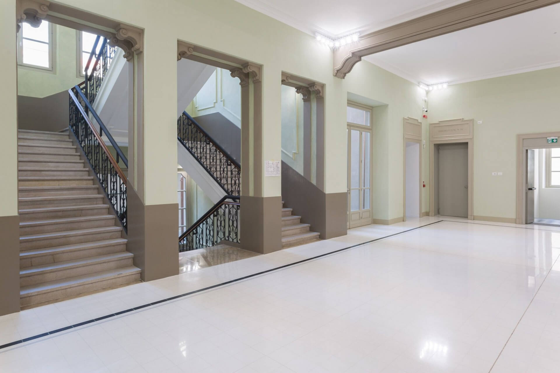 Interior distribution space in a restored historic building, featuring a large foyer with polished light-colored flooring and neutral-toned plastered walls with classic cornices and moldings. The interior façade is punctuated by a sequence of segmental arched openings with decorative trim, framing the monumental staircase, divided into multiple flights. The stairs are made of stone with ornamental wrought-iron parapets and wooden handrails, original features preserved and enhanced in the renovation project. Alongside the main space are glass windows and an integrated elevator shaft, demonstrating the incorporation of contemporary systems within the historic structure. Discreet linear lighting positioned along the upper perimeter emphasizes the moldings and contributes to a uniform reading of the space, highlighting the dialogue between traditional architectural elements and functional adaptations.