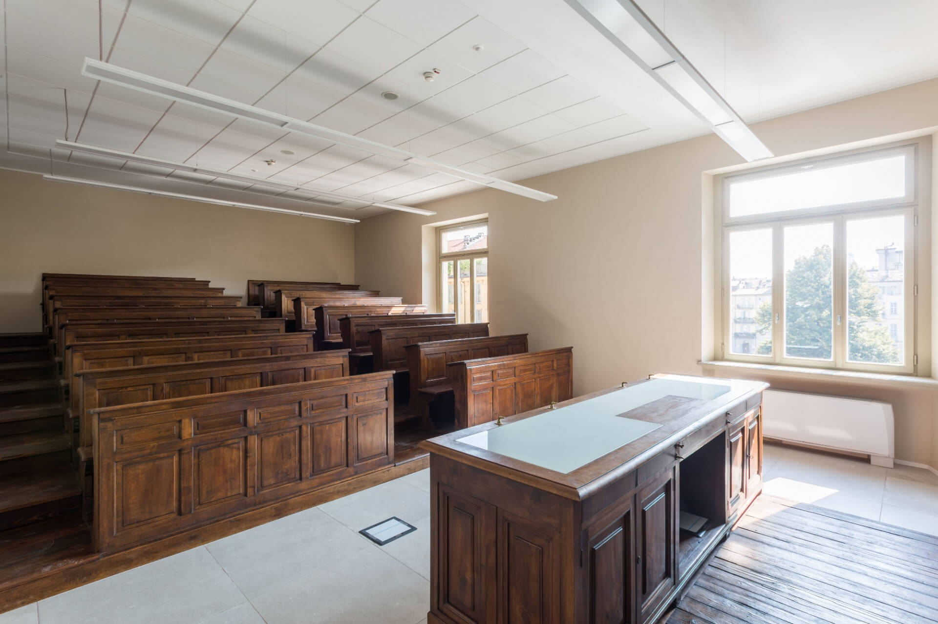 Classroom with a historic layout featuring wooden benches arranged on tiers, crafted from solid wood with molded paneling, facing a frontal wooden teacher's desk with an integrated worktop. The space is defined by neutral-toned plastered walls and large side windows that provide natural lighting, overlooking the urban surroundings outside. The ceiling features a modular false ceiling with suspended linear lighting fixtures and integrated technical devices, highlighting a contemporary intervention aimed at functionally adapting the space. The floor combines stone slab surfaces in the front area and wooden planks in the tiers, emphasizing the hierarchy of the spaces. The overall effect is that of a historic classroom repurposed for teaching purposes, where the original furnishings and layout are preserved and integrated with modern systems.