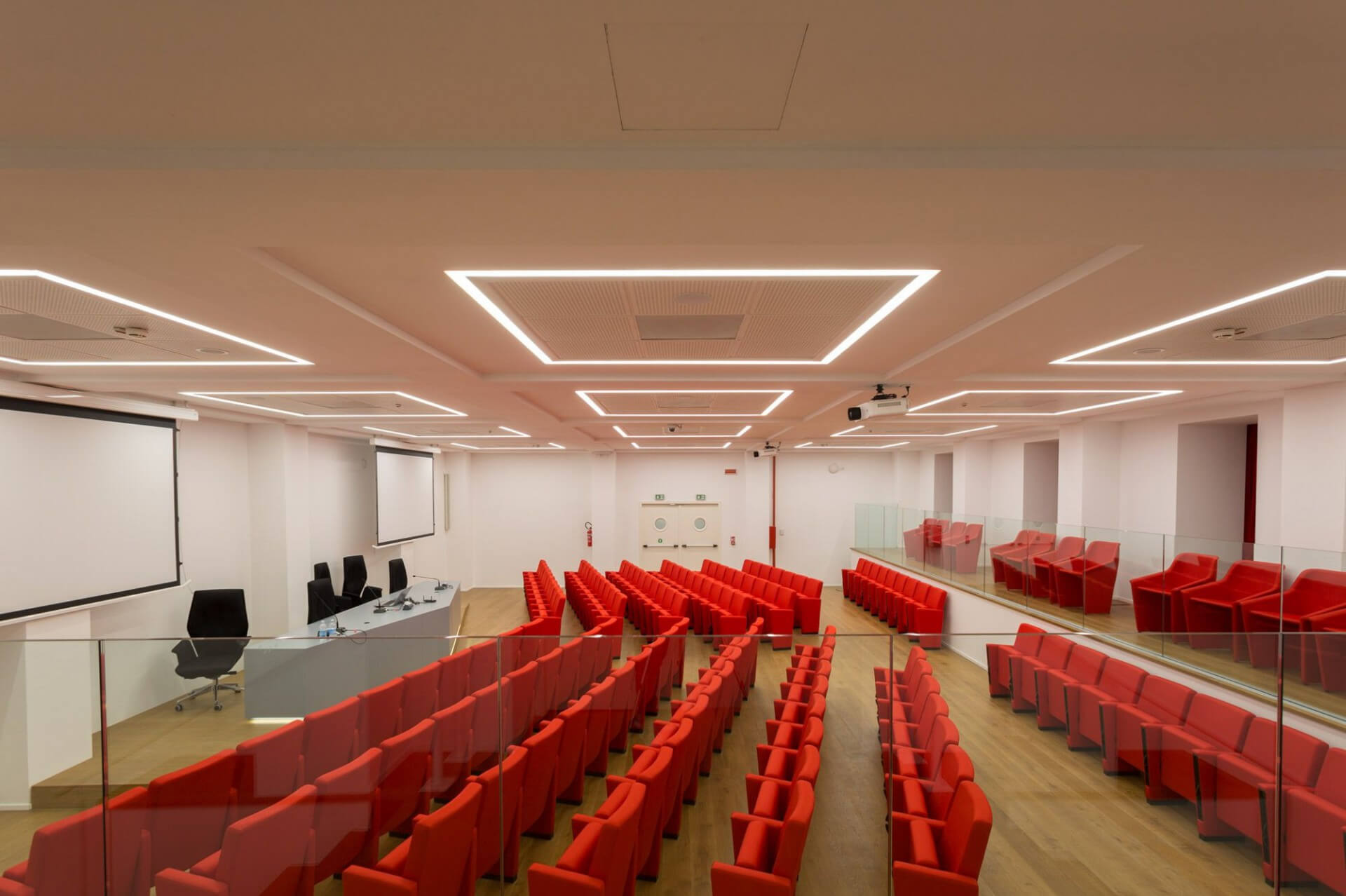 A contemporary conference room with an auditorium layout, featuring neat rows of red padded seats oriented toward the front stage, which houses a speaker table and projection screen. The space features a longitudinal configuration with wooden flooring and lateral glass parapets delimiting the raised areas. The ceiling is defined by a technical false ceiling with modular panels and LED lighting integrated into recessed geometric profiles, creating a regular and uniform light grid. The white plastered walls house projection screens and audiovisual equipment, highlighting the space's intended use for conferences, lectures, or events. The overall design expresses a contemporary architectural language, with attention to acoustics, visibility, and system integration, through the coordinated use of light surfaces, glass elements, and colorful furnishings that define the spatial hierarchy.