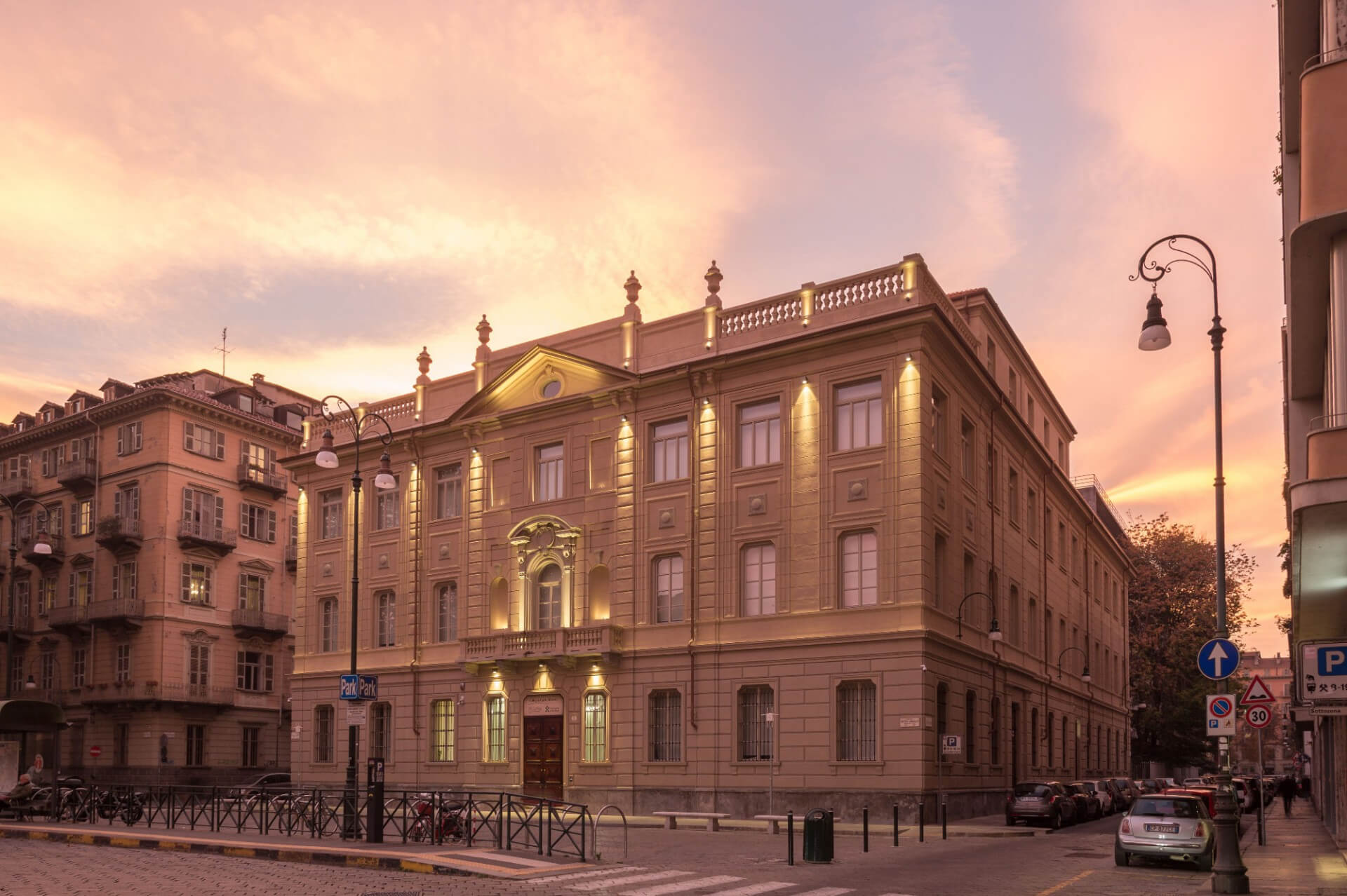 A neoclassical townhouse located on the corner of two streets, it features a symmetrical masonry façade with a rusticated base and upper levels punctuated by pilasters and string courses. The main façade features a monumental central entrance with a decorated portal, a balcony above, and a triangular pediment, flanked by regular windows with molded cornices. The roof is topped by a balustrade with ornamental elements. Warm architectural lighting emphasizes the sculptural details of the façade at dusk, while the surrounding urban context includes adjacent historic buildings, street lamps, signage, and cars parked along the roadway. The sunset sky, with its pink and orange hues, enhances the building's volumes and proportions.