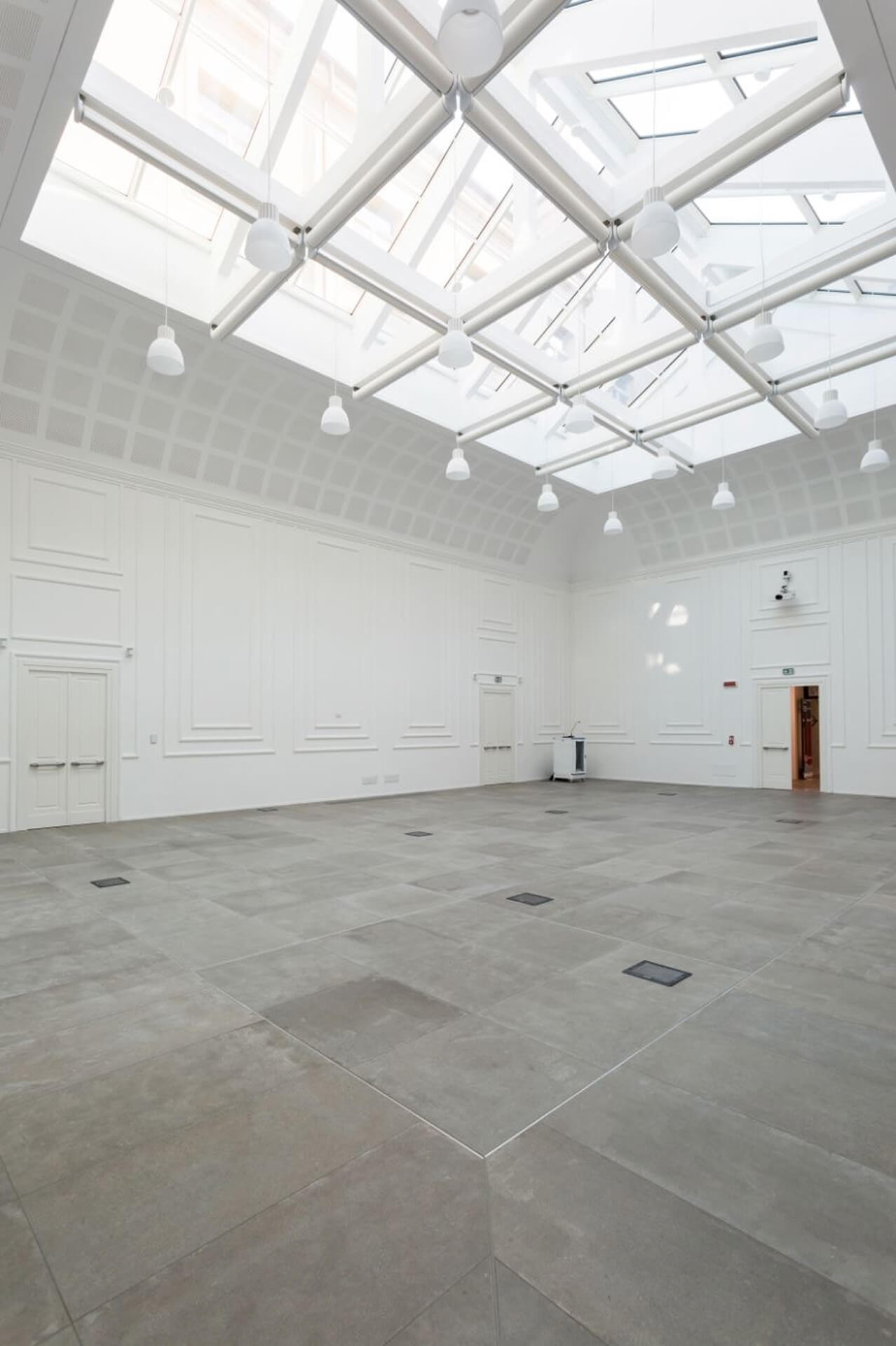 A large interior room with a restored historic shell and a contemporary glass roof, featuring a structural skylight with a modular metal lattice and exposed cylindrical nodes supporting transparent panels. Natural overhead light evenly illuminates the space, highlighting the white perimeter walls with preserved classical cornices and moldings. The floor is made of large, evenly laid gray stone slabs, integrated with flush-fitting technical elements. The glass ceiling is complemented by suspended lighting fixtures distributed throughout the structural grid. The unfurnished space highlights the contrast between the historic architectural language and the contemporary intervention, suggesting a space intended for exhibitions or conferences.