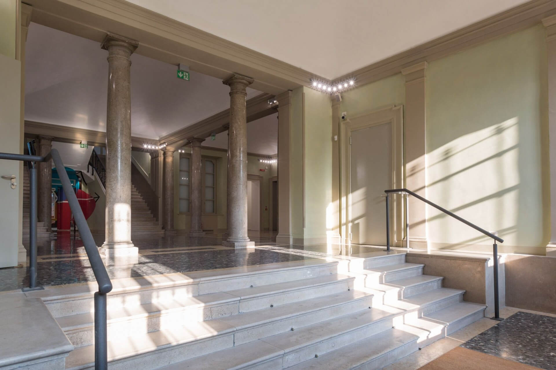 The interior entrance hall features a monumental layout, featuring a sequence of stone columns with simple capitals that define an internal portico and lead to a main staircase in the background. The space features light-colored plastered walls with classic cornices and moldings, complemented by contemporary touches such as linear metal handrails and a continuous LED lighting system positioned along the upper cornices. La pavimentazione combina superfici in pietra levigata e inserti lapidei più scuri, mentre una breve scalinata centrale articola i dislivelli tra gli spazi. La luce naturale radente, proveniente lateralmente, proietta ombre nette sulle superfici verticali e sui gradini, enfatizzando la plasticità degli elementi architettonici. L’insieme evidenzia un intervento di riqualificazione che mette in dialogo il linguaggio storico dell’edificio con soluzioni contemporanee, mantenendo la leggibilità dell’impianto originario.
