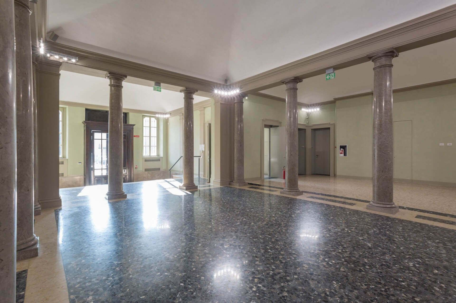 The interior space features a symmetrical layout punctuated by a sequence of polished stone columns supporting continuous architraves, defining a sort of internal portico open on multiple sides. The space features dark terrazzo flooring with a reflective finish, framed by lighter perimeter bands that highlight the geometry of the space. Le pareti intonacate in tonalità neutre presentano cornici e modanature classiche semplificate, integrate da interventi contemporanei quali illuminazione lineare a LED posizionata lungo il perimetro superiore e corpi illuminanti direzionali. Sullo sfondo si leggono aperture vetrate e ingressi secondari che articolano i percorsi interni, mentre la luce naturale proveniente dall’ingresso principale contribuisce a enfatizzare la profondità dello spazio e la matericità delle superfici lapidee. L’insieme restituisce un ambiente di rappresentanza riqualificato, in cui elementi strutturali e decorativi storici dialogano con soluzioni tecnologiche contemporanee, mantenendo una chiara leggibilità dell’impianto originario.