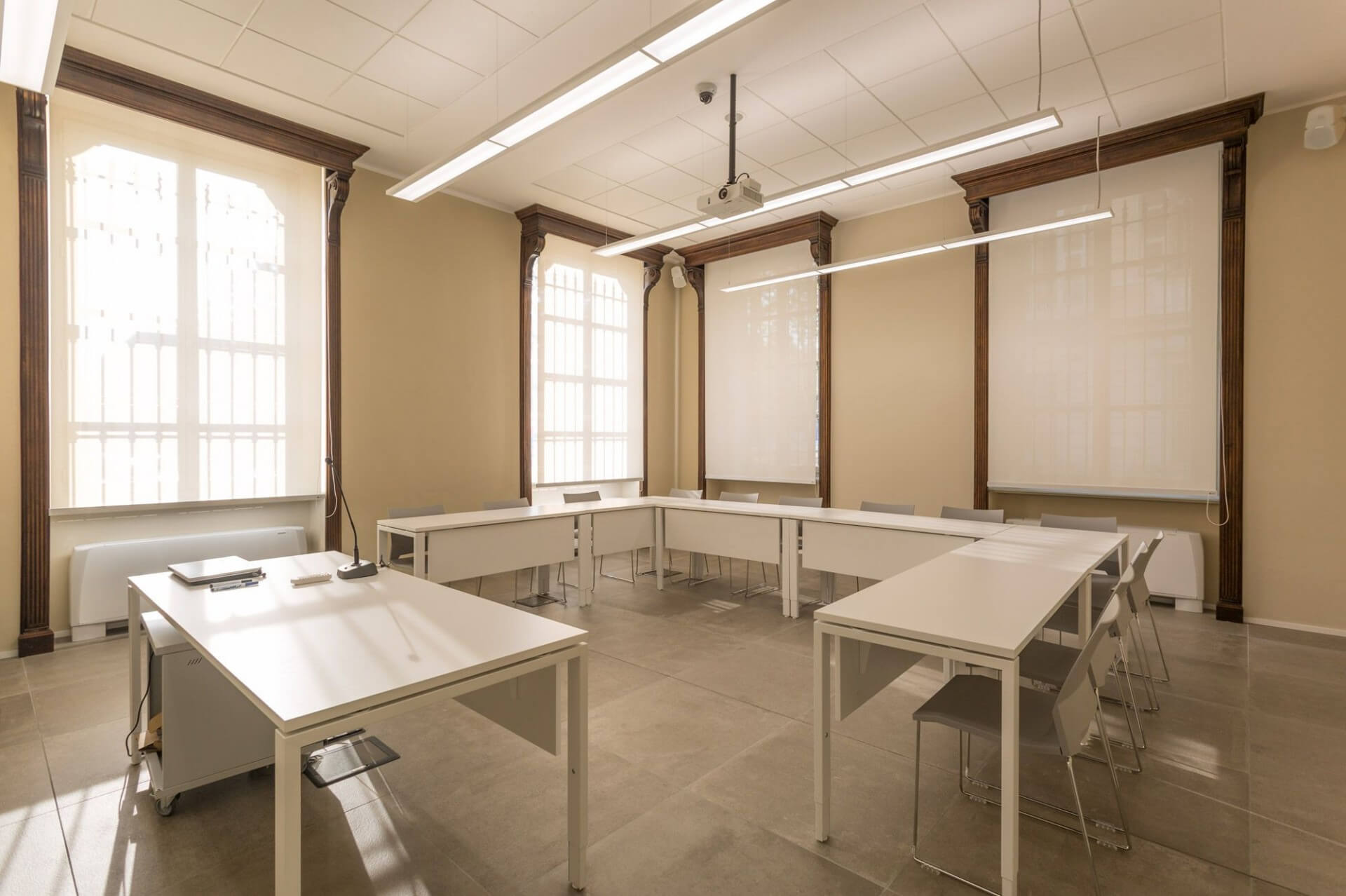 A multipurpose classroom created in a renovated historic building, featuring tall vertical windows with original frames and molded wooden frames framing the openings. The space features neutral-toned plastered walls and a modular suspended ceiling with integrated linear lighting fixtures, from which a central projector for presentations is suspended. L’arredo contemporaneo è composto da tavoli modulari bianchi disposti a configurazione a U, con sedute leggere coordinate, suggerendo un uso didattico o per riunioni. Il pavimento in grandi lastre ceramiche a finitura opaca contribuisce alla percezione ordinata e funzionale dello spazio. La luce naturale filtrata dalle schermature alle finestre si combina con l’illuminazione artificiale, evidenziando il dialogo tra elementi storici conservati e interventi tecnologici contemporanei.