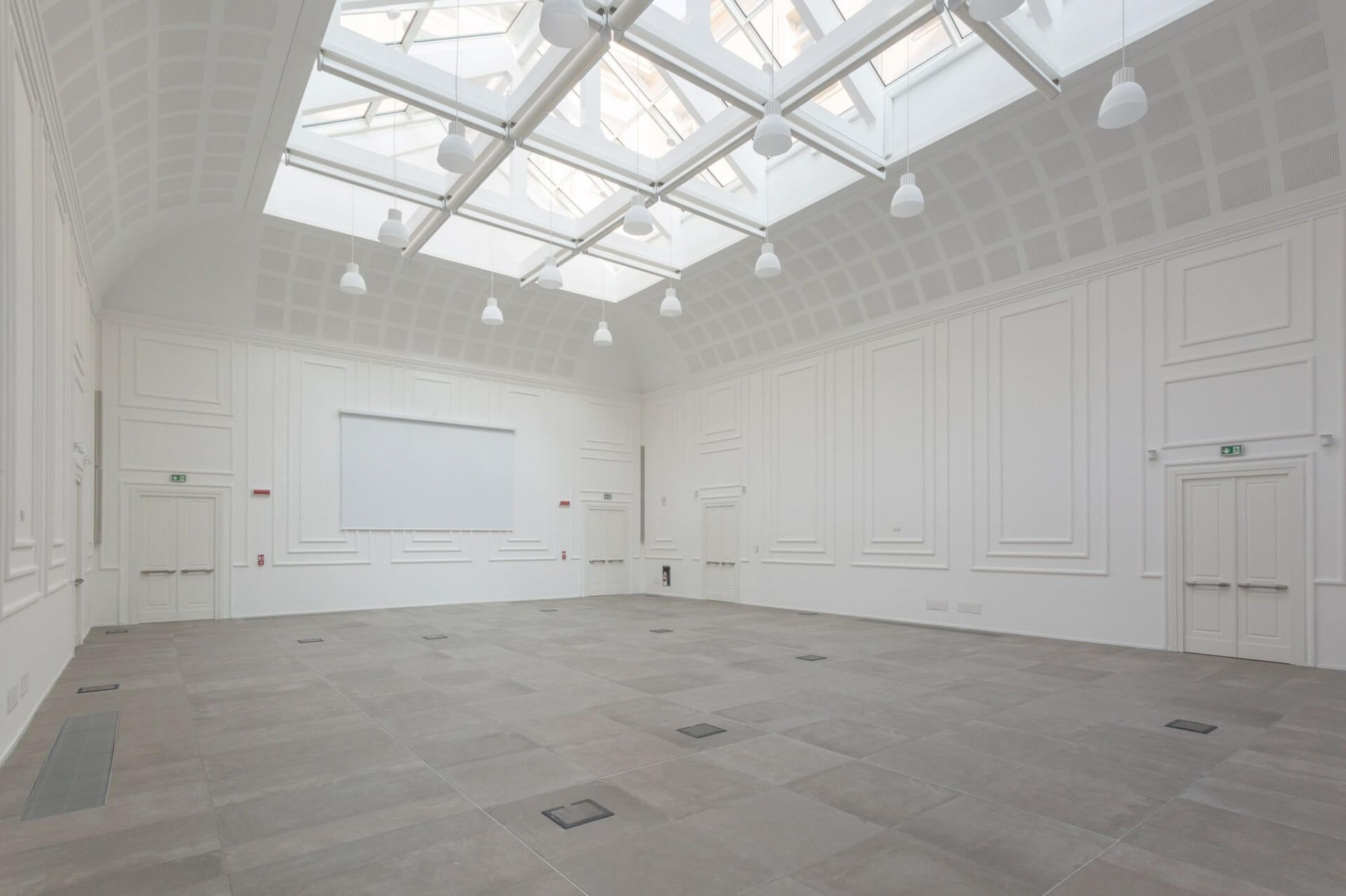 A large double-height room with a restored historic shell and a contemporary glass roof, featuring a structural skylight with a modular metal lattice and exposed nodes supporting transparent panels. Diffused natural light from above evenly illuminates the space, highlighting the white perimeter walls articulated by classic cornices and frames. Il pavimento è realizzato in grandi lastre lapidee grigie con integrazione di elementi tecnici a filo, mentre il sistema di illuminazione sospesa segue la maglia strutturale della copertura. Su una delle pareti principali è integrato uno schermo per proiezioni, suggerendo una destinazione d’uso per eventi, conferenze o attività espositive. L’insieme evidenzia il dialogo tra linguaggio storico e intervento contemporaneo, con particolare attenzione alla luce e alla leggibilità spaziale.