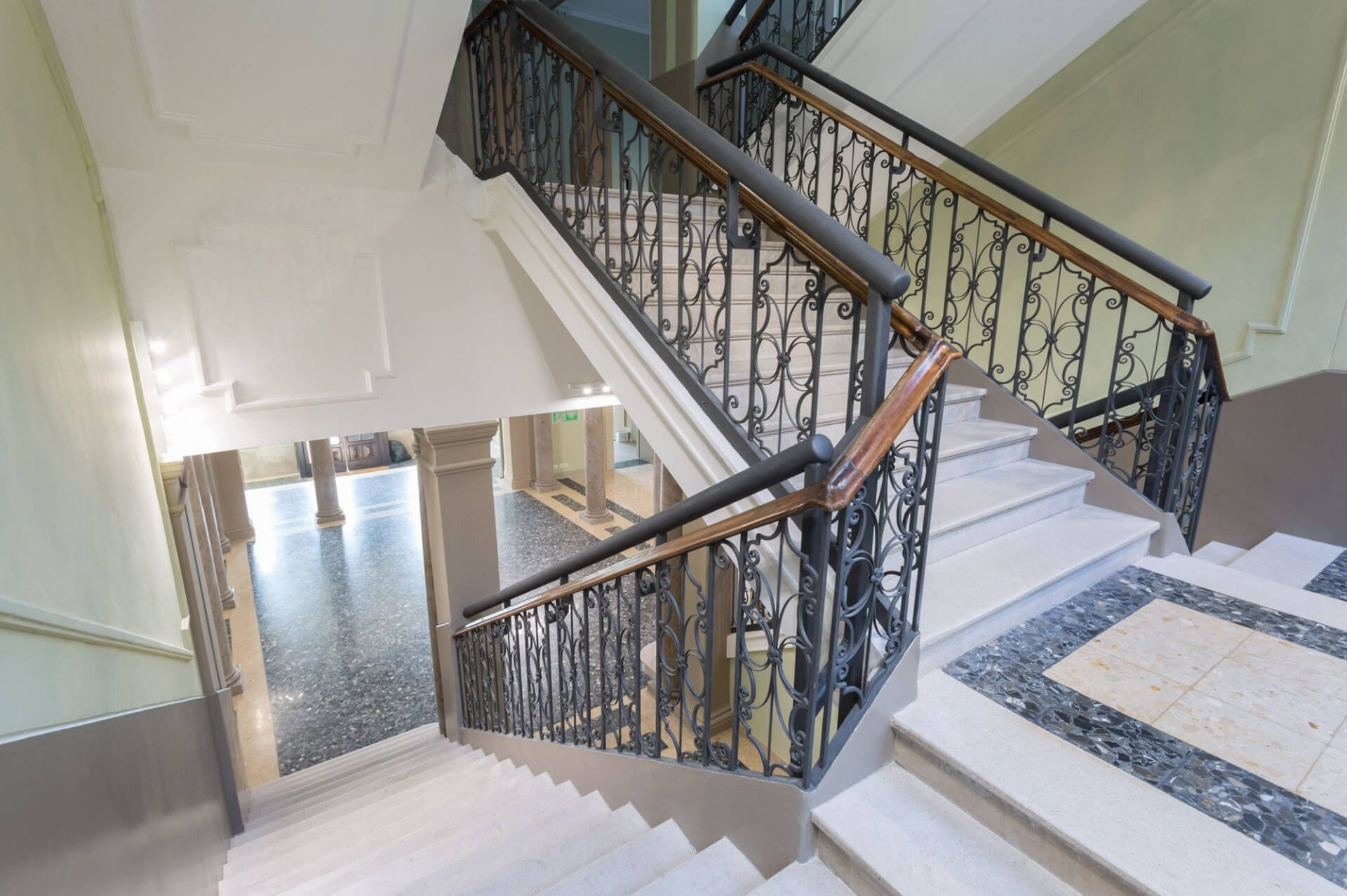 Monumental internal staircase in a restored historic building, featuring light-colored stone ramps with terrazzo-clad landings and decorative stone inserts. The wrought iron parapet features a continuous ornamental design and a wooden handrail, an original feature preserved and integrated into the renovation project. La scala si sviluppa attorno a un vano aperto che mette in relazione i diversi livelli dell’edificio, con affaccio sullo spazio distributivo sottostante scandito da colonne in pietra. Le pareti intonacate in tonalità neutre presentano cornici e modanature classiche semplificate, mentre l’illuminazione contemporanea discreta valorizza i dettagli architettonici senza alterare la lettura storica dell’insieme. L’immagine evidenzia il dialogo tra elementi costruttivi originali e interventi di adeguamento funzionale, con particolare attenzione alla conservazione delle finiture e alla continuità materica tra pavimentazioni, strutture verticali e parapetti.