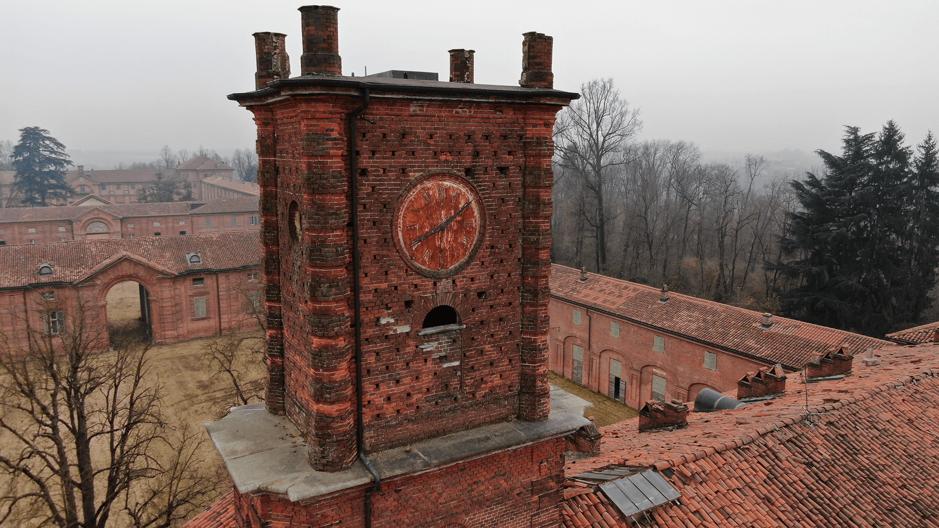Torre in mattoni rossi con grande orologio circolare sulla facciata, che emerge sopra i tetti in coppi di un complesso storico; sullo sfondo alberi spogli e altri edifici della cittadella sotto un cielo grigio e nebbioso.