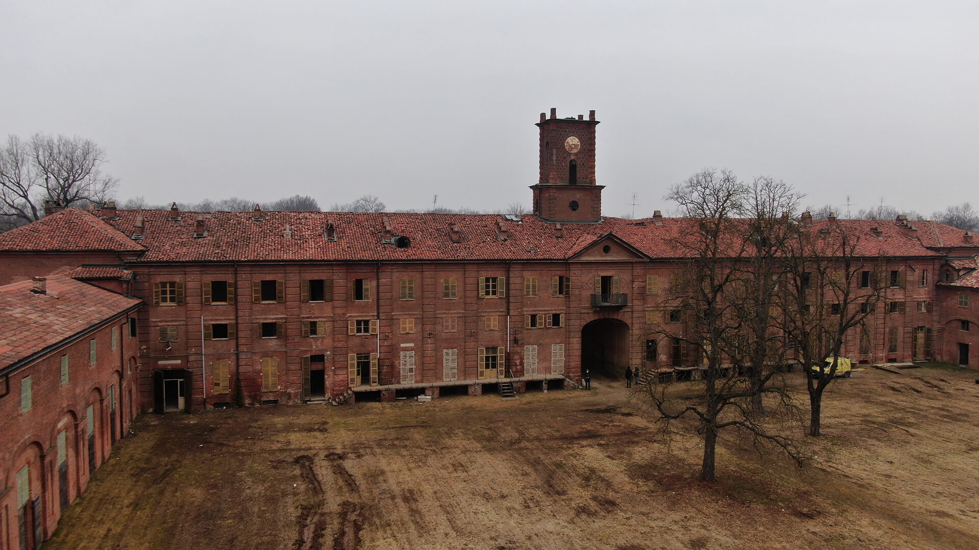 Grande edificio storico in mattoni rossi con tetto in coppi e torre centrale con orologio; davanti si apre un ampio cortile con terreno spoglio e alberi senza foglie, sotto un cielo grigio e nebbioso.