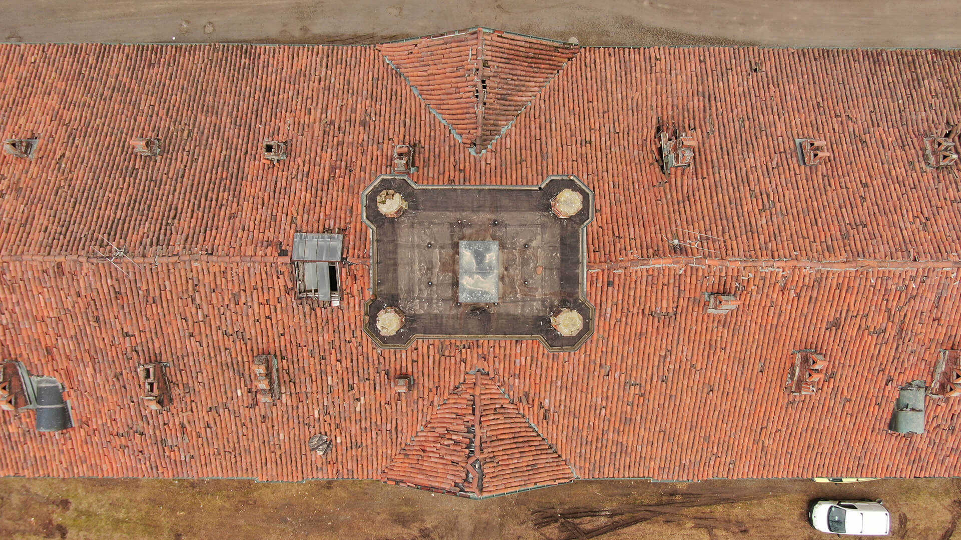 Vista dall’alto di un ampio tetto in coppi rossi con struttura centrale sopraelevata e piccola apertura vetrata; intorno sono visibili lucernari, comignoli e segni di usura delle tegole, con una porzione di terreno e un’auto parcheggiata ai margini.
