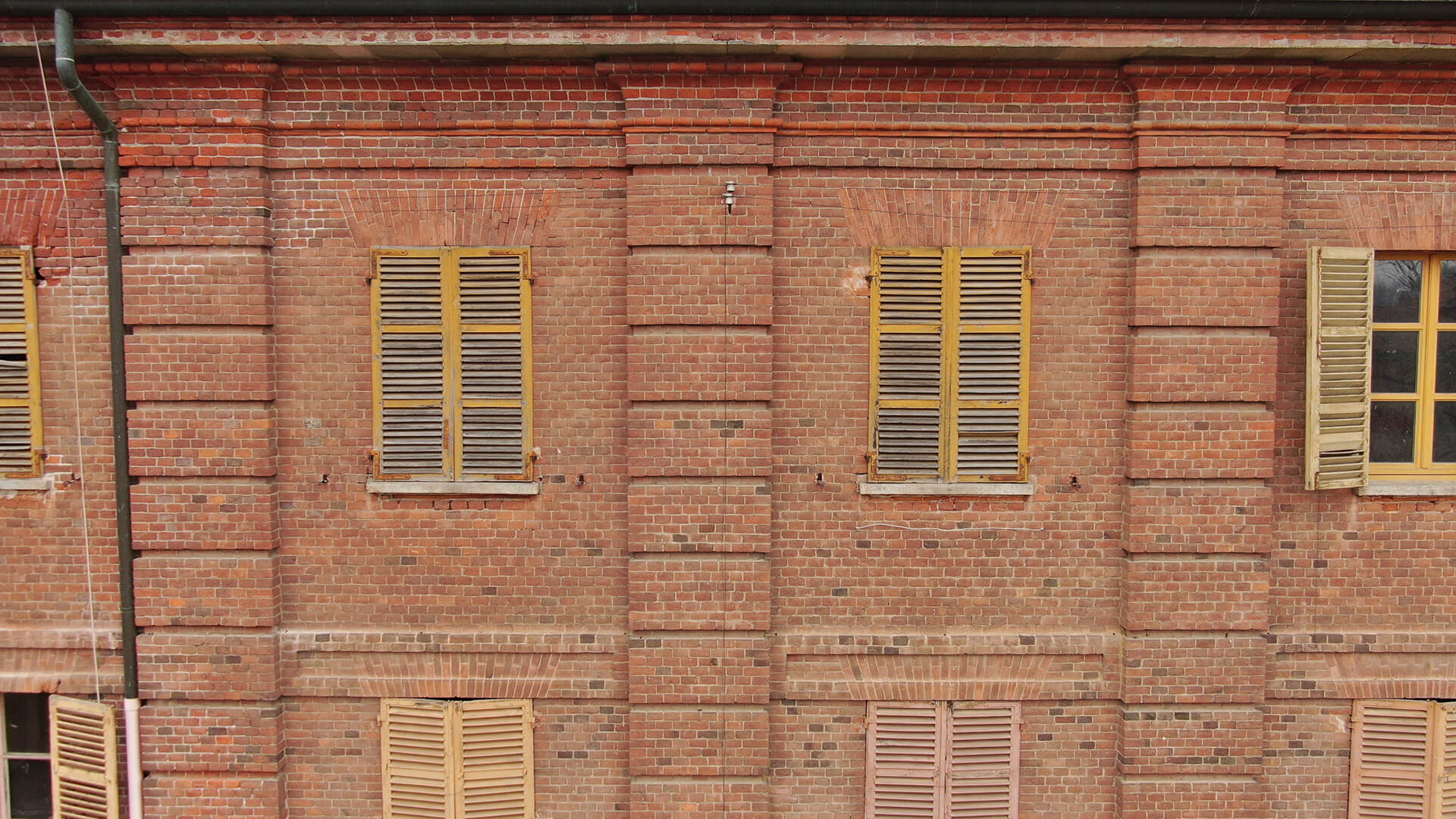 Facciata di edificio storico in mattoni rossi con finestre rettangolari e persiane in legno color ocra, disposte in modo regolare lungo il muro.