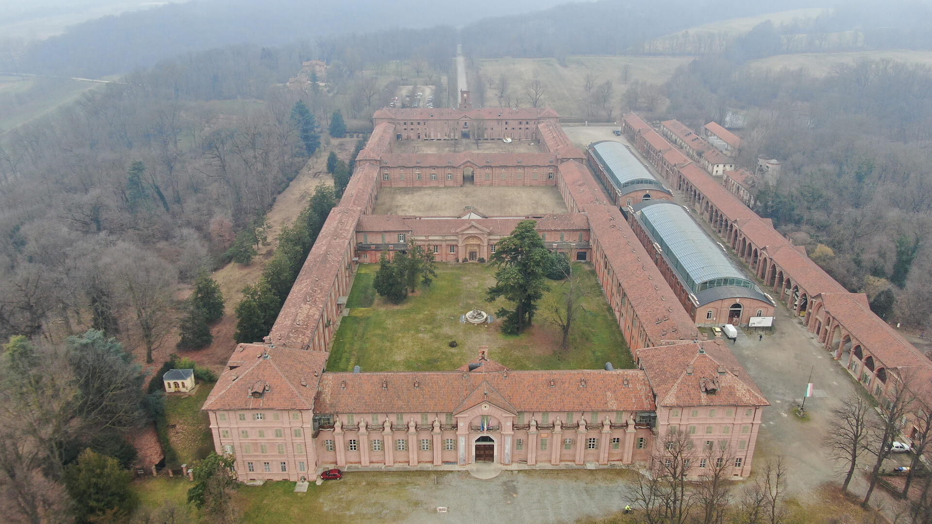 Vista aerea di un grande complesso storico in mattoni con tetti in coppi disposti attorno a un ampio cortile centrale alberato; l’edificio principale con facciata monumentale si trova in primo piano, mentre intorno si estende un paesaggio di alberi spogli e campagna avvolta nella nebbia.