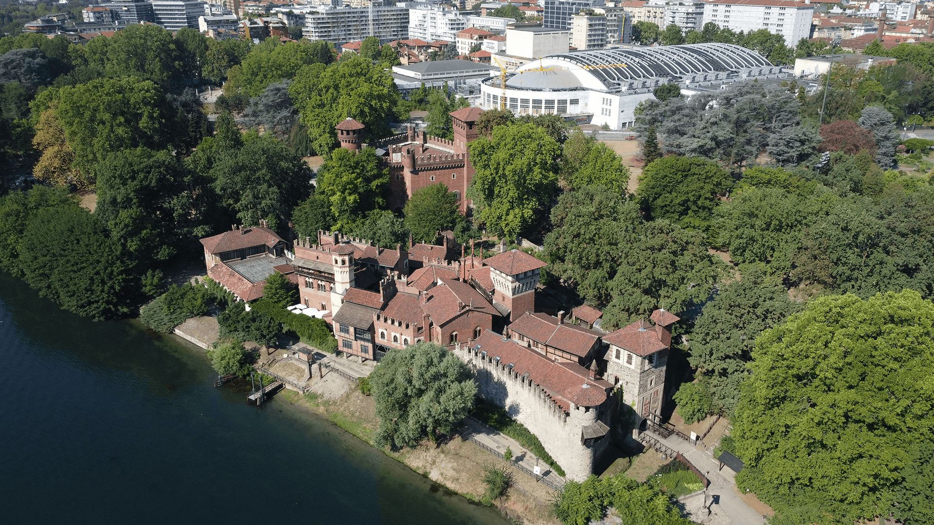 Vista aerea di un complesso storico in mattoni con torri e mura merlate, circondato da alberi e affacciato su uno specchio d’acqua, con edifici urbani e strutture moderne sullo sfondo.