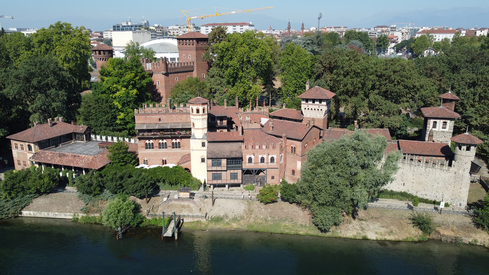 Close-up aerial view of a historic architectural complex with the characteristics of a fortified residence, composed of brick buildings with towers, battlements, and tile roofs, located along the bank of a stream and surrounded by a large, wooded park. The building system features buildings arranged around internal courtyards and perimeter walls, with defensive elements and volumes from different eras integrated into a single whole. In the background, contemporary urban buildings, sports facilities, and construction cranes are visible, highlighting the relationship between the restored historic complex and the surrounding urban context.
