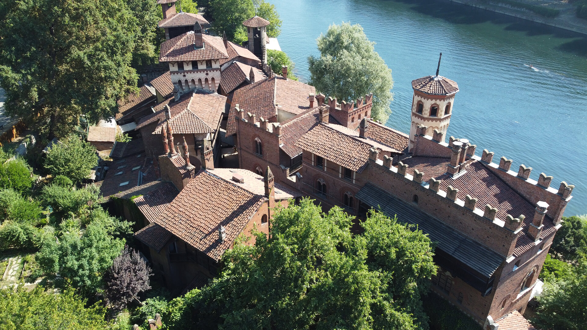 Close-up aerial view of a historic brick complex with neo-medieval architectural features, composed of complex volumes with cylindrical towers, battlements, loggias, and tiled roofs, located along the bank of a stream and surrounded by dense vegetation. The composition shows a succession of interconnected buildings, with fortified perimeter walls and internal courtyards, highlighting the complex's unified layout. The presence of the river and the surrounding park emphasizes the landscape connection between the restored historic architecture and the natural environment.