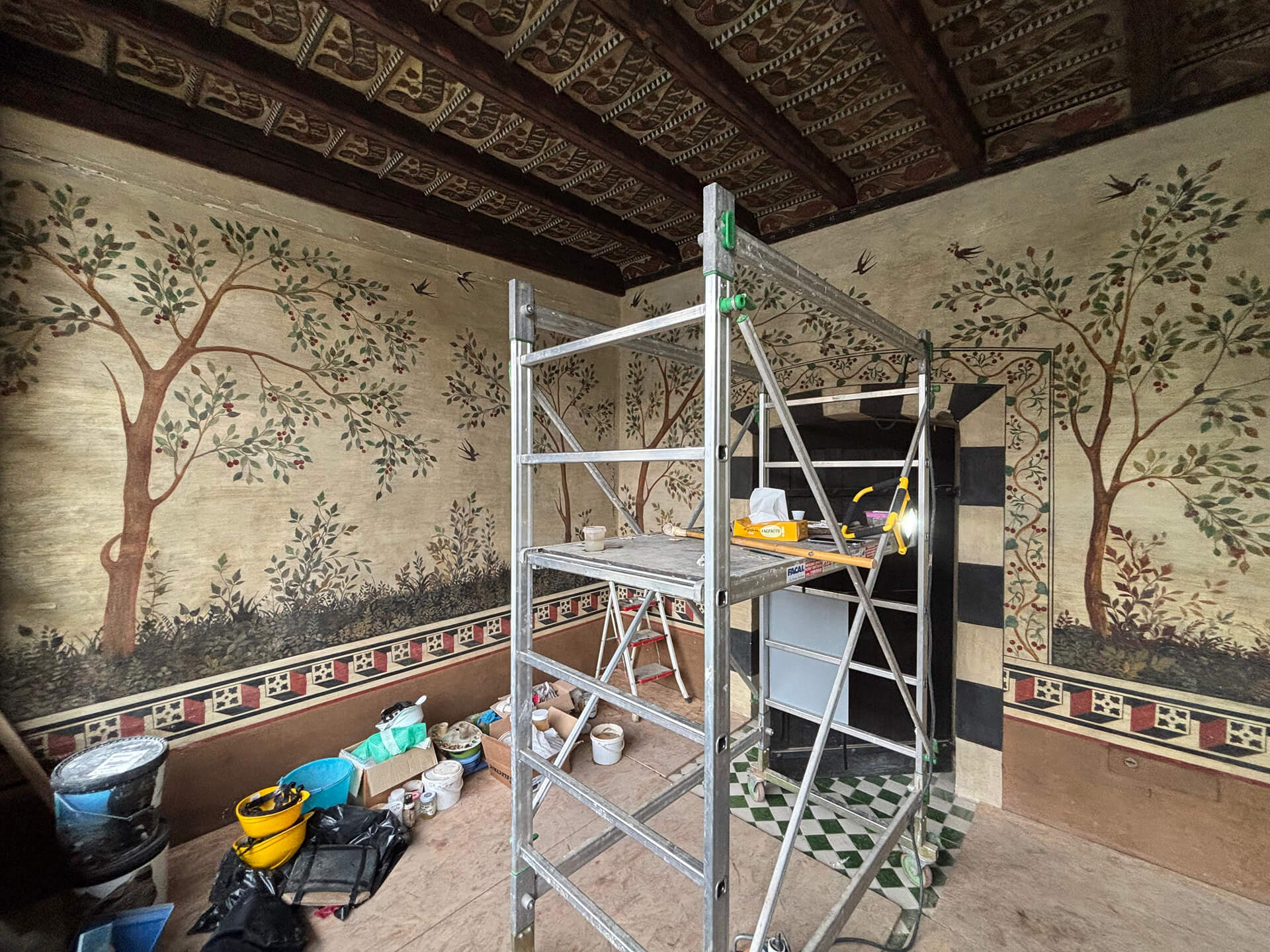 Interior of a historic building during restoration, featuring walls decorated with murals depicting trees, plants, and birds on a light background, with a geometric ornamental band below. The wooden beamed ceiling with decorated coffers is fully preserved and features polychrome painted motifs. At the center of the room is metal scaffolding with construction equipment, buckets, and restoration materials used for cleaning and consolidating the decorated surfaces. The scene documents a phase of conservation work in a historic setting with original decorative features.
