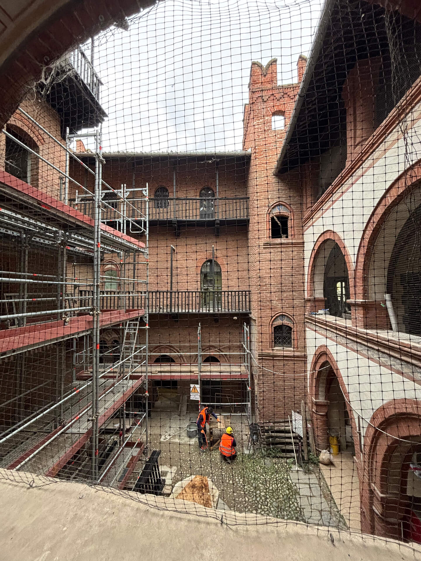 The internal courtyard of a historic brick complex during restoration, featuring exposed brick façades with arches, loggias, and a crenellated turret. Metal scaffolding is present on multiple levels for consolidation and maintenance work on the walls and balconies. A construction site safety net is visible in the foreground, while workers wearing safety gear are working on the ground. The image documents a phase of work within the courtyard, highlighting the original architectural structure and the ongoing restoration work.