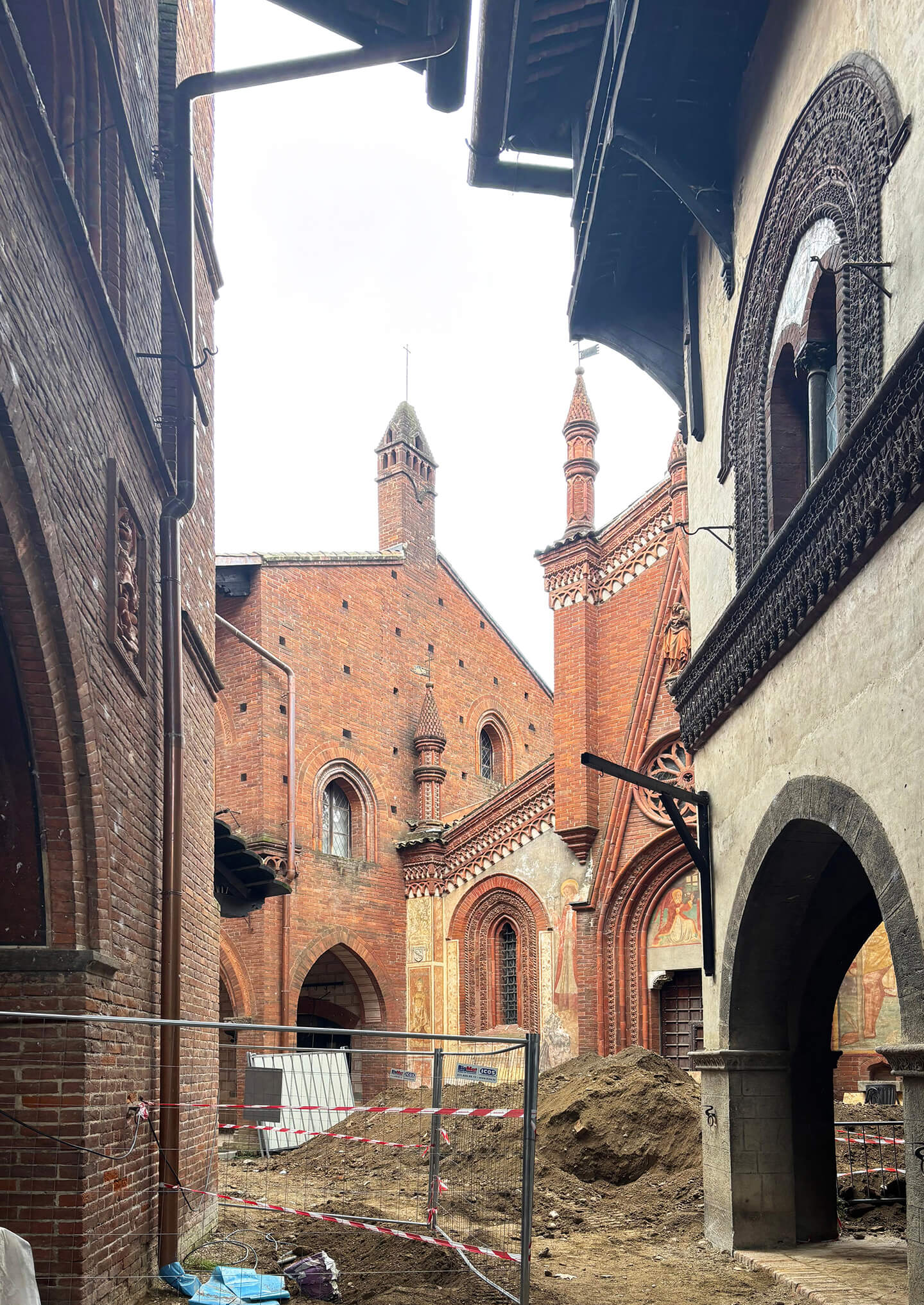 View of the internal courtyard of a historic brick complex during restoration, characterized by neo-Gothic architecture with pointed arches, terracotta decorations, and crenellated turrets. The facades feature decorative elements, frescoes, and molded brick cornices, while in the center of the courtyard, excavations and mounds of earth are visible, enclosed by construction site safety fencing. The image shows a work phase with a restricted area and temporary structures, highlighting the original architectural context and the restoration work underway within the historic complex.