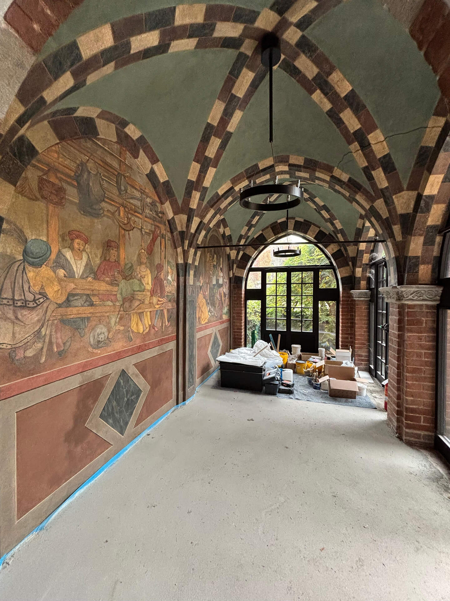 Vaulted corridor of a historic building during restoration, featuring cross vaults with alternating two-tone bands and frescoed walls depicting figurative scenes from historical periods. Brick walls and columns with sculpted capitals support the arches of the loggia, while at the end is a glass door leading to the external courtyard. Visible on the floor are materials, equipment, and construction containers used for cleaning and consolidating the decorated surfaces, documenting a phase of conservative restoration of the pictorial and architectural elements.