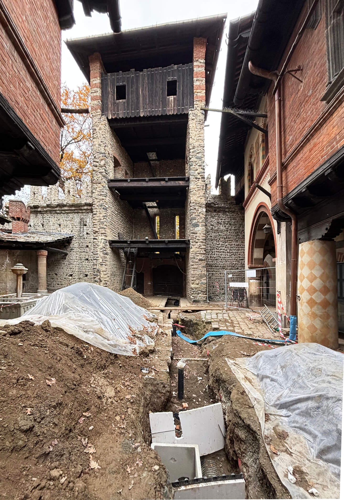 The internal courtyard of a historic complex undergoing restoration, featuring mixed stone and brick masonry structures, featuring a tower with wooden balconies and decorated porticoes. The central area is undergoing archaeological excavations and plant engineering work, with open trenches, pipes, and mounds of earth protected by tarps. Security fencing, temporary stairs, and consolidation work are visible, while the facades feature painted decorative elements, brick columns, and arches. The image documents a phase of ongoing work within the courtyard, highlighting the relationship between the historic structures and the restoration and renovation interventions.
