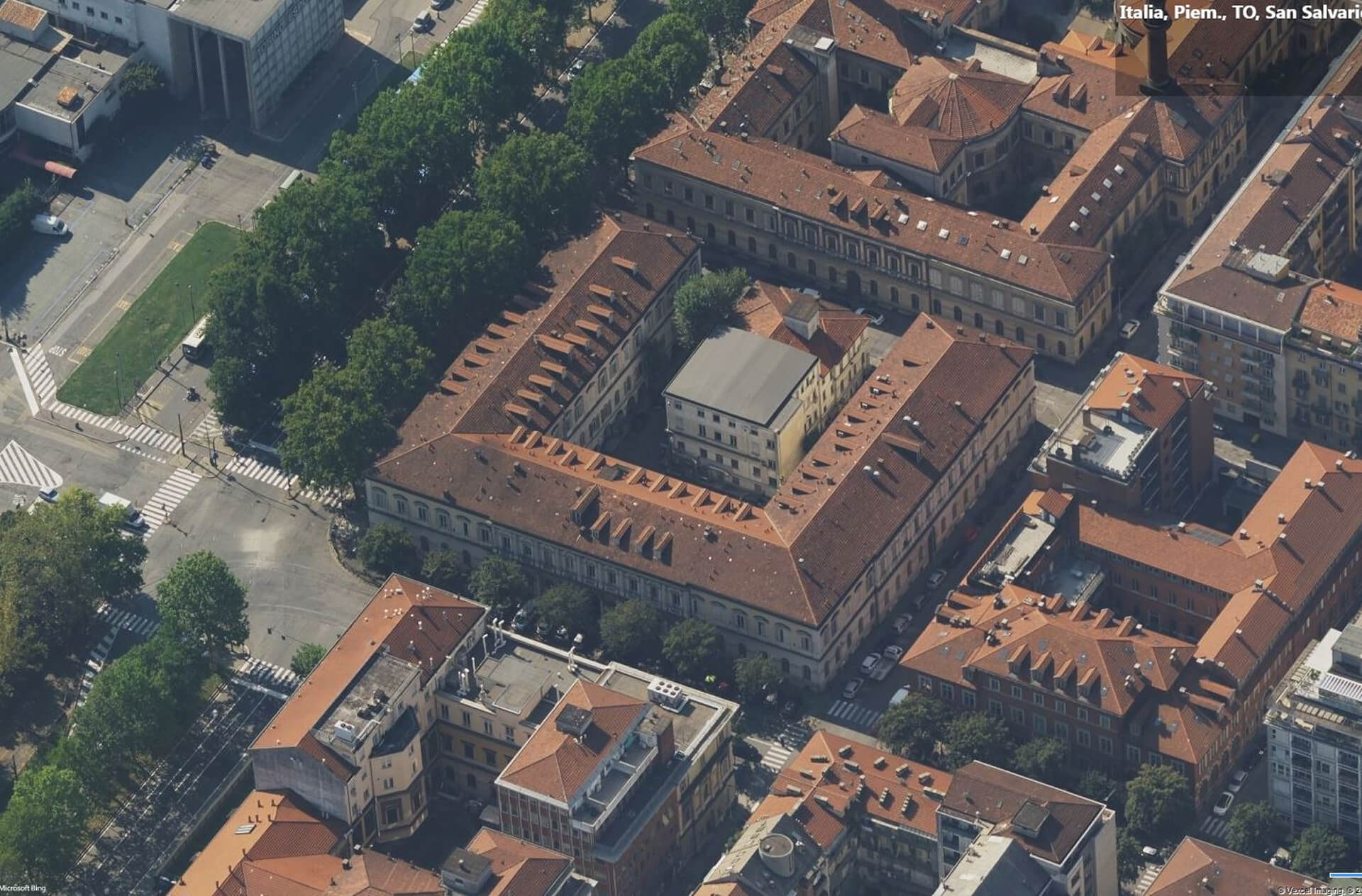 Vista aerea del tessuto urbano con isolato storico a corte chiusa caratterizzato da edifici con coperture in laterizio e impianto rettangolare organizzato attorno a cortile interno. Il complesso, inserito in un contesto urbano compatto con strade alberate e incroci viari, è circondato da edifici residenziali e strutture pubbliche di diversa epoca. L’immagine evidenzia la morfologia dell’isolato, la continuità delle facciate lungo il perimetro e il rapporto tra il complesso architettonico e il sistema urbano circostante.