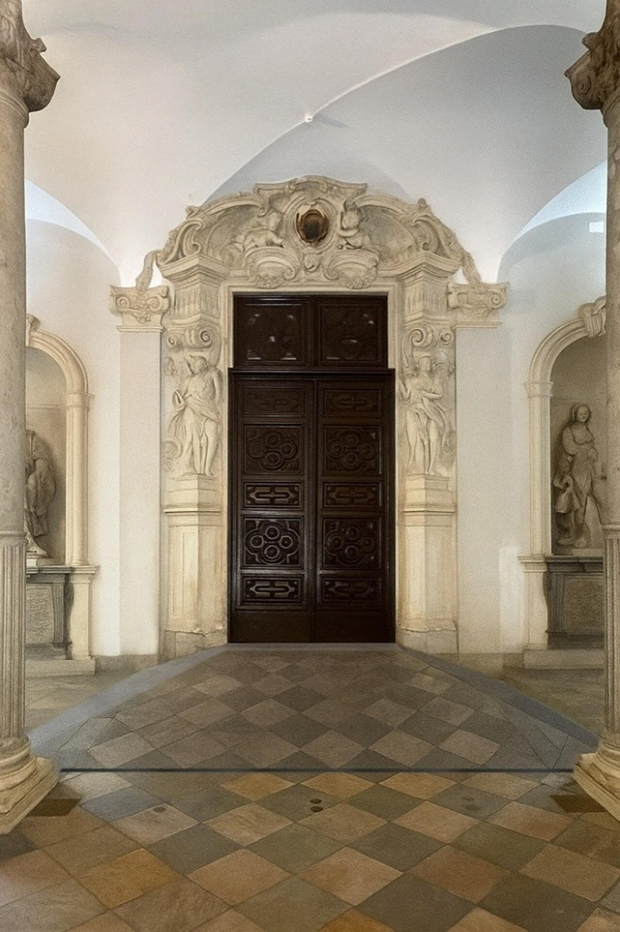 L'immagine mostra un ambiente ricco di dettagli architettonici barocchi. Ecco alcune caratteristiche chiave: Porta ornamentale: La porta in legno scuro al centro è finemente decorata con un design geometrico, che richiama l'arte barocca. La cornice della porta è altrettanto decorata con sculture e motivi floreali, un tratto distintivo dello stile. Architettura barocca: Le colonne e gli archi, insieme alla stuccatura dettagliata, sono tipici del periodo barocco. Le sculture nelle nicchie laterali sembrano rappresentare figure umane, anch'esse con un tocco di eleganza barocca, raffigurando probabilmente figure allegoriche o mitologiche. Pavimentazione in pietra: Il pavimento è caratterizzato da piastrelle disposte in un motivo a rombi, con diverse tonalità di pietra che creano un effetto visivo raffinato. Illuminazione e spaziosità: L'illuminazione naturale proveniente dalle finestre non visibili nell'immagine contribuisce a evidenziare le texture e i dettagli architettonici, dando un'atmosfera serena e maestosa. Questa immagine cattura la bellezza e la grandiosità tipiche di un edificio progettato in stile barocco, dove ogni elemento architettonico ha una forte componente decorativa e simbolica.