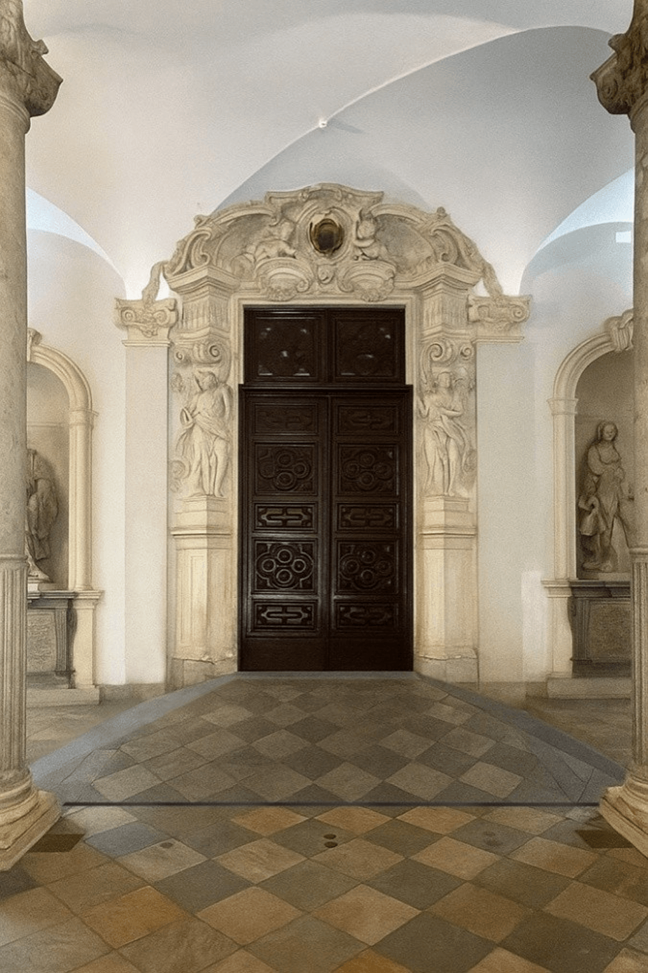 L'immagine mostra un ambiente ricco di dettagli architettonici barocchi. Ecco alcune caratteristiche chiave: Porta ornamentale: La porta in legno scuro al centro è finemente decorata con un design geometrico, che richiama l'arte barocca. La cornice della porta è altrettanto decorata con sculture e motivi floreali, un tratto distintivo dello stile. Architettura barocca: Le colonne e gli archi, insieme alla stuccatura dettagliata, sono tipici del periodo barocco. Le sculture nelle nicchie laterali sembrano rappresentare figure umane, anch'esse con un tocco di eleganza barocca, raffigurando probabilmente figure allegoriche o mitologiche. Pavimentazione in pietra: Il pavimento è caratterizzato da piastrelle disposte in un motivo a rombi, con diverse tonalità di pietra che creano un effetto visivo raffinato. Illuminazione e spaziosità: L'illuminazione naturale proveniente dalle finestre non visibili nell'immagine contribuisce a evidenziare le texture e i dettagli architettonici, dando un'atmosfera serena e maestosa. Questa immagine cattura la bellezza e la grandiosità tipiche di un edificio progettato in stile barocco, dove ogni elemento architettonico ha una forte componente decorativa e simbolica.