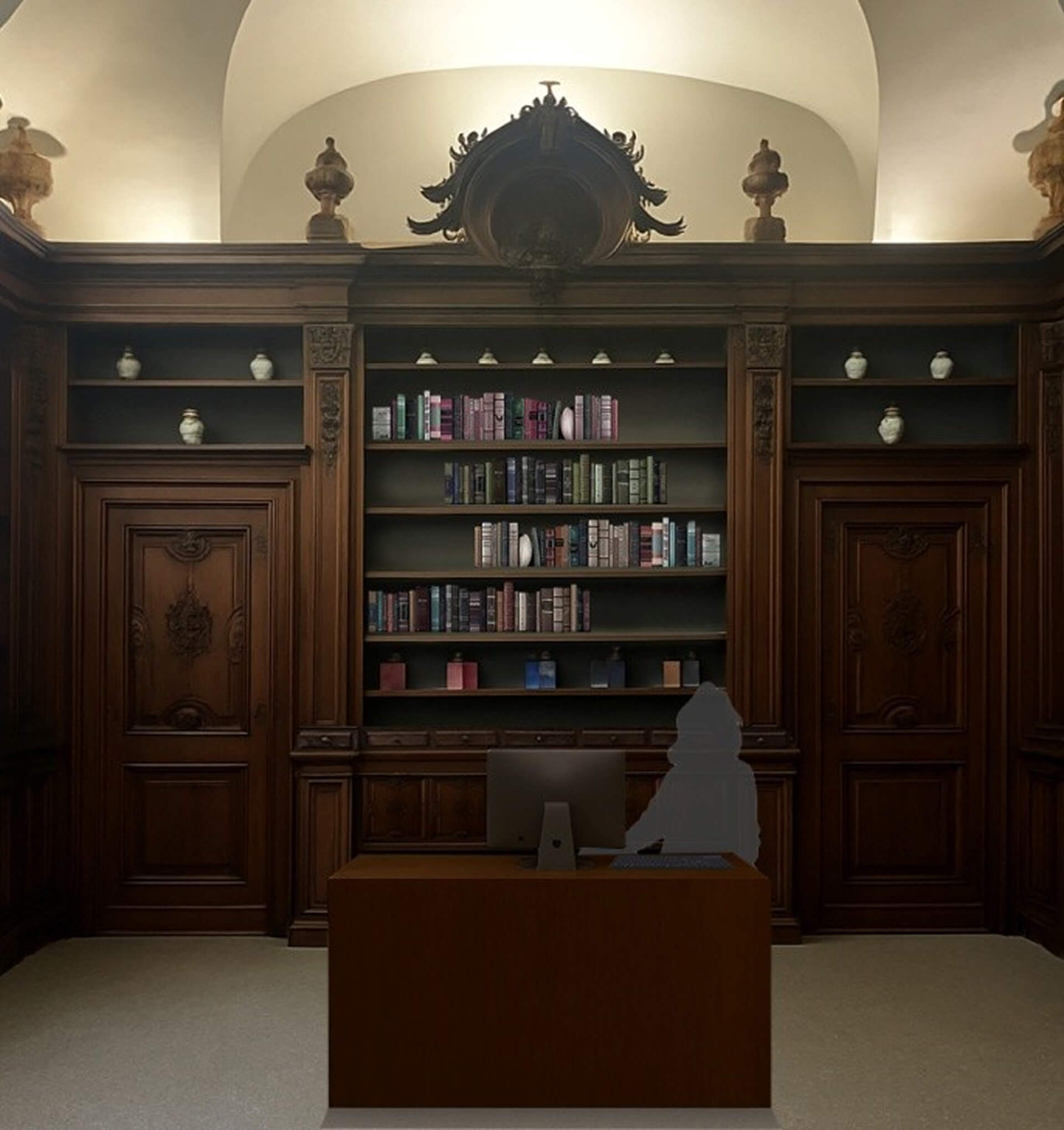 L'immagine mostra un elegante studio o ufficio, che evoca un'atmosfera storica e raffinata. Ecco alcuni dettagli notevoli: Mobili in legno scuro: Le librerie e le porte in legno massiccio, con intagli ornamentali e finiture dettagliate, indicano un design classico e ricercato, probabilmente ispirato allo stile barocco o rococò. Questi elementi contribuiscono a un'aria di nobiltà e prestigio. Librerie ricche di libri e decorazioni: Le mensole piene di libri ben disposti, con una varietà di colori che vanno dal blu al rosa, creano un contrasto elegante con il legno scuro. Anche gli oggetti decorativi, come le anfore sui ripiani superiori, aggiungono un tocco artistico. Soffitto elegante e illuminazione discreta: Il soffitto a volta con una finitura ricca e la luce che filtra da sopra suggeriscono un ambiente ampio e ben curato. L'illuminazione è delicata, ma crea una sensazione di calore e accoglienza. Scrivania moderna: Nonostante l'ambiente storico, la scrivania moderna con un computer e una sedia suggerisce che l'ambiente è destinato a un uso attivo, forse come ufficio per un professionista o uno studio privato. Questo contrasto tra antico e moderno crea una fusione interessante tra passato e presente. Atmosfera serena e funzionale: Le linee pulite e l'ordine delle librerie e degli oggetti decorativi conferiscono all'ambiente una sensazione di tranquillità e serenità, rendendolo perfetto per la lettura, lo studio o il lavoro. In generale, l'ambiente rappresenta una fusione elegante di elementi storici e moderni, creando un luogo perfetto per riflessione e lavoro intellettuale in un contesto ricco di tradizione.