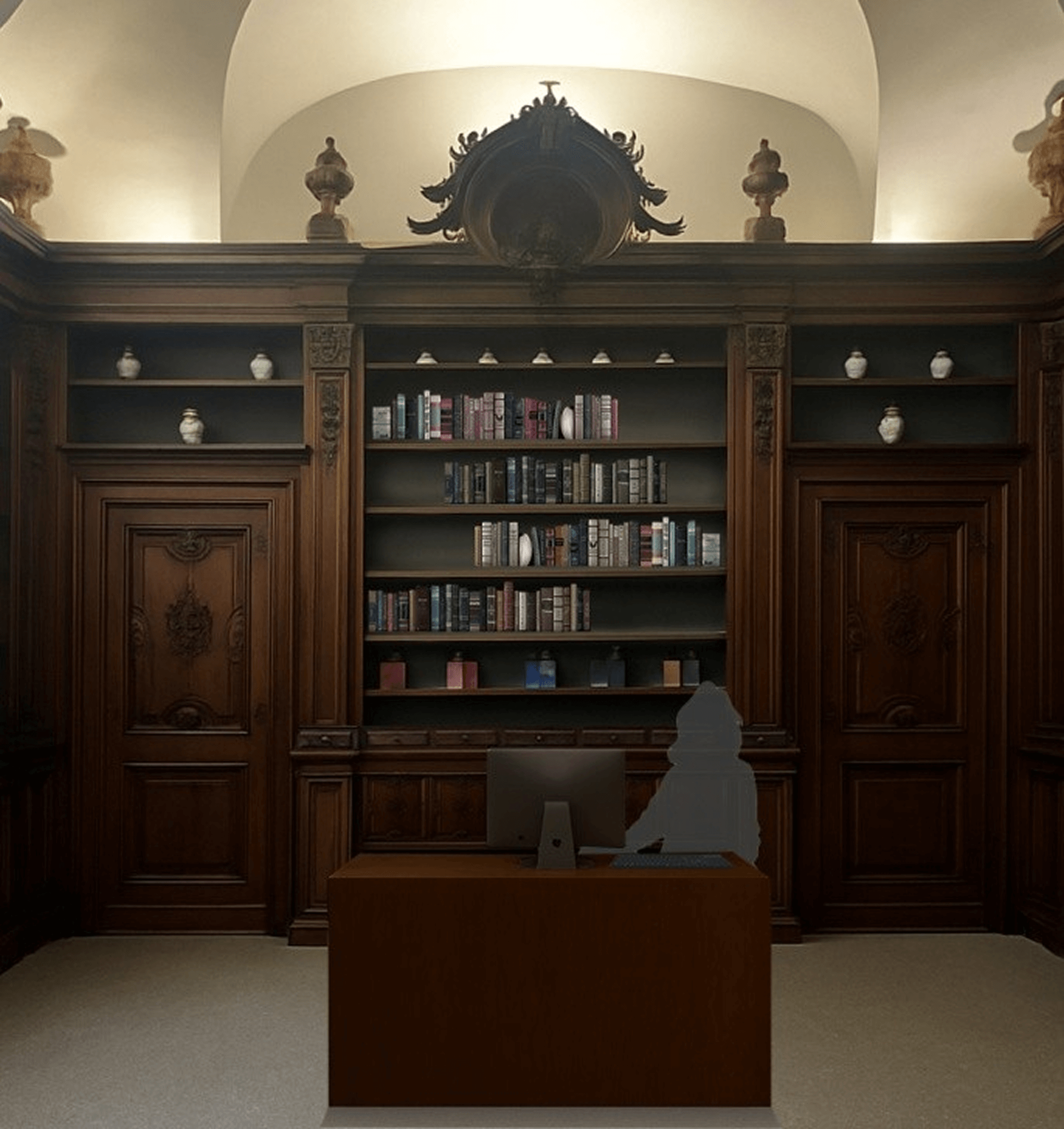 L'immagine mostra un elegante studio o ufficio, che evoca un'atmosfera storica e raffinata. Ecco alcuni dettagli notevoli: Mobili in legno scuro: Le librerie e le porte in legno massiccio, con intagli ornamentali e finiture dettagliate, indicano un design classico e ricercato, probabilmente ispirato allo stile barocco o rococò. Questi elementi contribuiscono a un'aria di nobiltà e prestigio. Librerie ricche di libri e decorazioni: Le mensole piene di libri ben disposti, con una varietà di colori che vanno dal blu al rosa, creano un contrasto elegante con il legno scuro. Anche gli oggetti decorativi, come le anfore sui ripiani superiori, aggiungono un tocco artistico. Soffitto elegante e illuminazione discreta: Il soffitto a volta con una finitura ricca e la luce che filtra da sopra suggeriscono un ambiente ampio e ben curato. L'illuminazione è delicata, ma crea una sensazione di calore e accoglienza. Scrivania moderna: Nonostante l'ambiente storico, la scrivania moderna con un computer e una sedia suggerisce che l'ambiente è destinato a un uso attivo, forse come ufficio per un professionista o uno studio privato. Questo contrasto tra antico e moderno crea una fusione interessante tra passato e presente. Atmosfera serena e funzionale: Le linee pulite e l'ordine delle librerie e degli oggetti decorativi conferiscono all'ambiente una sensazione di tranquillità e serenità, rendendolo perfetto per la lettura, lo studio o il lavoro. In generale, l'ambiente rappresenta una fusione elegante di elementi storici e moderni, creando un luogo perfetto per riflessione e lavoro intellettuale in un contesto ricco di tradizione.