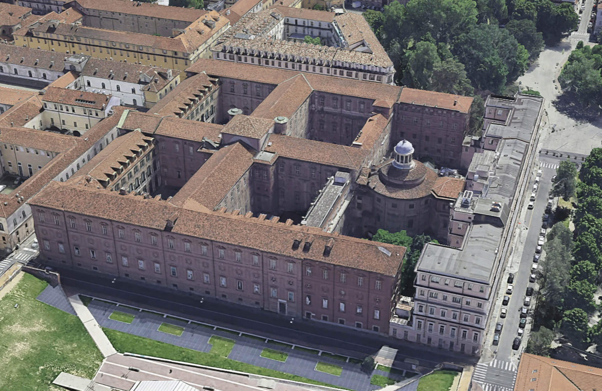 The image shows an aerial view of an architectural complex, which appears to be a large historic building or convent with a courtyard structure. Here are some significant details: Symmetrical complex: The main building is laid out symmetrically, with a central courtyard visible in the middle. The pitched terracotta roof is a distinctive element, suggesting classical architecture. Central dome: A dome can be seen in the central part of the complex, which could be a significant architectural feature, such as a church or main hall. The dome is often a sign of great architectural and functional importance in historic buildings. Restoration in progress: In some places, the shelving is visible, suggesting the building is undergoing restoration or maintenance. This type of restoration is common for historic buildings of this magnitude. Green belt and garden: Green areas can be seen around the building, contributing to a tranquil atmosphere and the feel of a residence or an academic/clerical institution. Long facades and elegant windows: The long facades with numerous windows could indicate that the building was intended to house many people, such as a convent, monastery, or other type of historical institution. This structure appears to have a rich history and sophisticated architectural design, suggesting it is a building of great cultural and historical value.