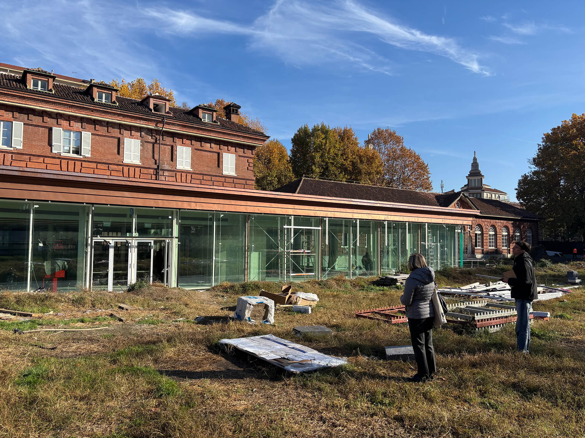 Edificio storico in mattoni rossi con tetto in coppi e persiane chiare, affiancato da una nuova struttura vetrata in fase di allestimento; nell’area antistante, prato con materiali da cantiere sparsi e due persone in piedi che osservano i lavori sotto un cielo sereno.