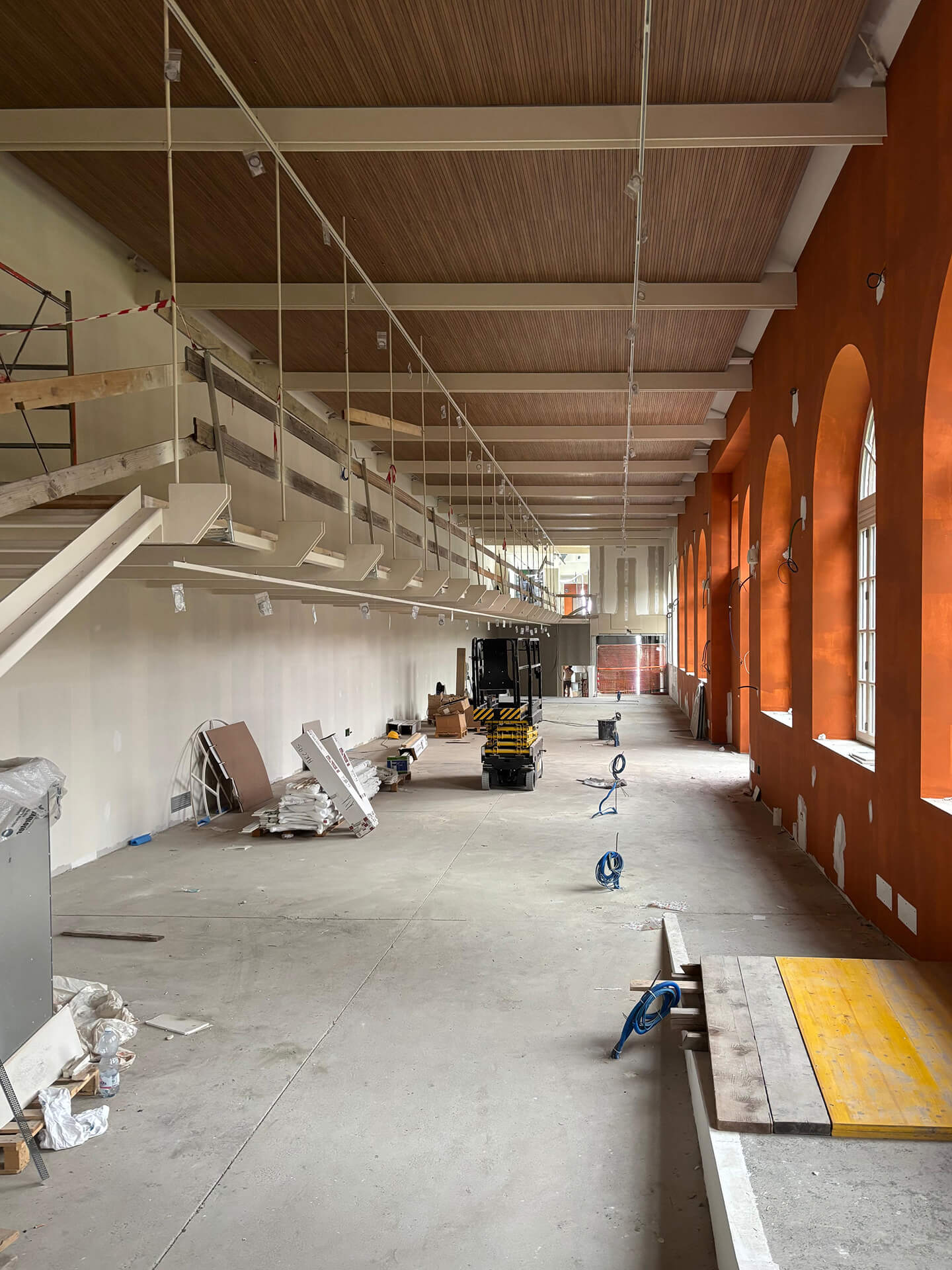 Ampio ambiente interno in fase di ristrutturazione, con soffitto in legno a travi bianche, parete destra arancione con finestre ad arco e parete sinistra bianca con mensole sospese; sul pavimento sono presenti materiali, cavi e attrezzature da cantiere, tra cui una piattaforma elevatrice al centro della sala.