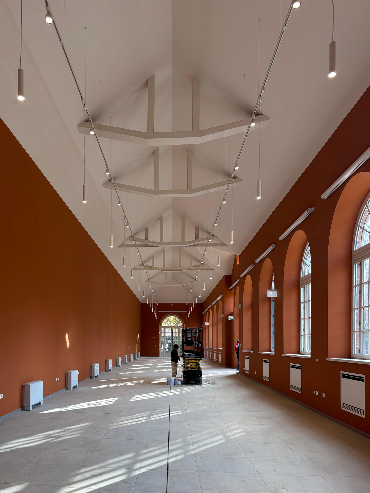 Lungo salone restaurato con pareti color terracotta e alte finestre ad arco sulla destra; soffitto bianco con travi a vista e file di luci sospese, pavimento chiaro illuminato dal sole e, al centro, una piattaforma elevatrice con un operatore al lavoro.