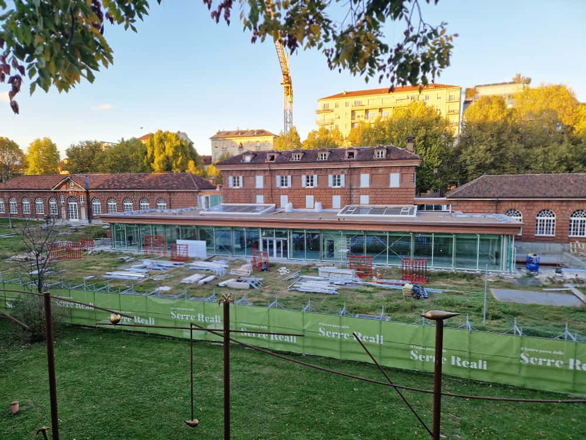 Edificio storico in mattoni rossi con ampie finestre ad arco e una nuova struttura vetrata in fase di completamento sul lato frontale; nell’area del cantiere sono visibili materiali da costruzione e recinzioni verdi con la scritta “Serre Reali”, sullo sfondo alberi e palazzi illuminati dalla luce del tramonto.