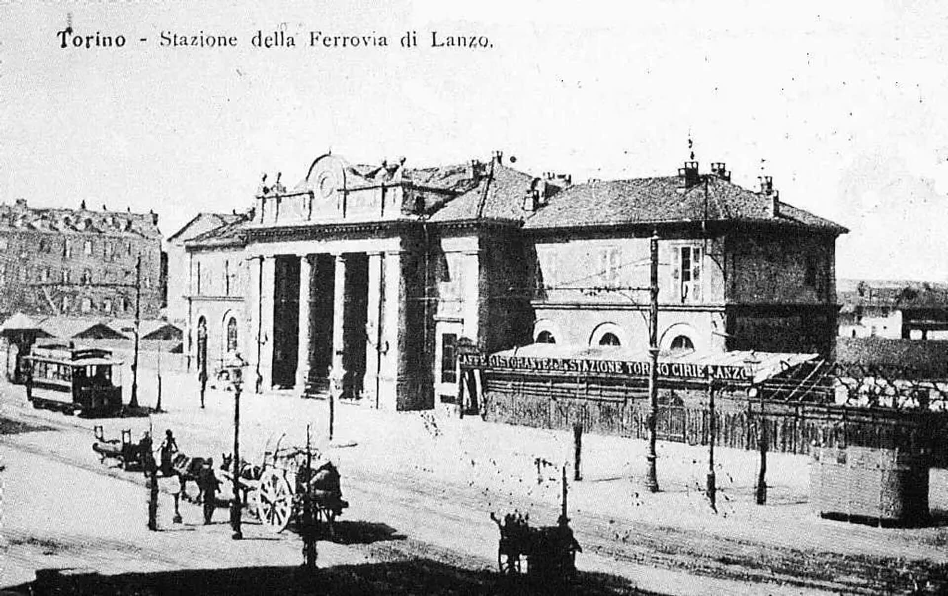 Fotografia storica in bianco e nero della Stazione della Ferrovia di Lanzo a Torino, risalente ai primi anni del XX secolo, che documenta l’aspetto originario dell’edificio prima delle trasformazioni successive. L’immagine mostra il prospetto principale dell’edificio ferroviario, caratterizzato da una composizione simmetrica con corpo centrale monumentale e ali laterali più basse, impostate su un volume compatto con copertura a falde. L’ingresso principale è evidenziato da un portico con colonne di ordine classico che sorreggono un frontone decorato, elemento architettonico che conferisce all’edificio un carattere rappresentativo e istituzionale, tipico delle architetture ferroviarie di fine Ottocento. Le aperture ad arco al piano terra e le finestre regolari al piano superiore definiscono una facciata ordinata e ritmata, costruita secondo criteri compositivi accademici. In primo piano è visibile lo spazio urbano antistante la stazione, con strada sterrata, carrozze trainate da cavalli, lampioni e alcune persone in sosta, elementi che testimoniano il contesto urbano dell’epoca e il ruolo della stazione come nodo di scambio tra città e ferrovia. Sul lato sinistro compare un tram, mentre sullo sfondo si intravedono edifici residenziali e strutture di servizio legate all’infrastruttura ferroviaria. La fotografia ha valore documentario e consente di riconoscere i caratteri originari dell’edificio storico oggetto di intervento, mettendo in evidenza la configurazione architettonica iniziale, la relazione con lo spazio pubblico e l’importanza della stazione nel sistema dei trasporti torinese.